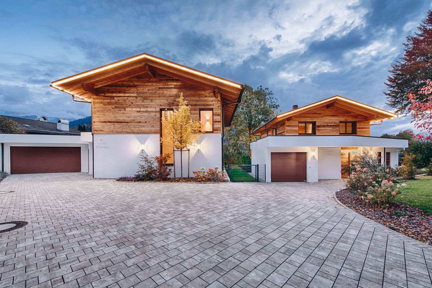 Modern luxury homes with wooden accents and white exterior featuring curved paver driveway at twilight.