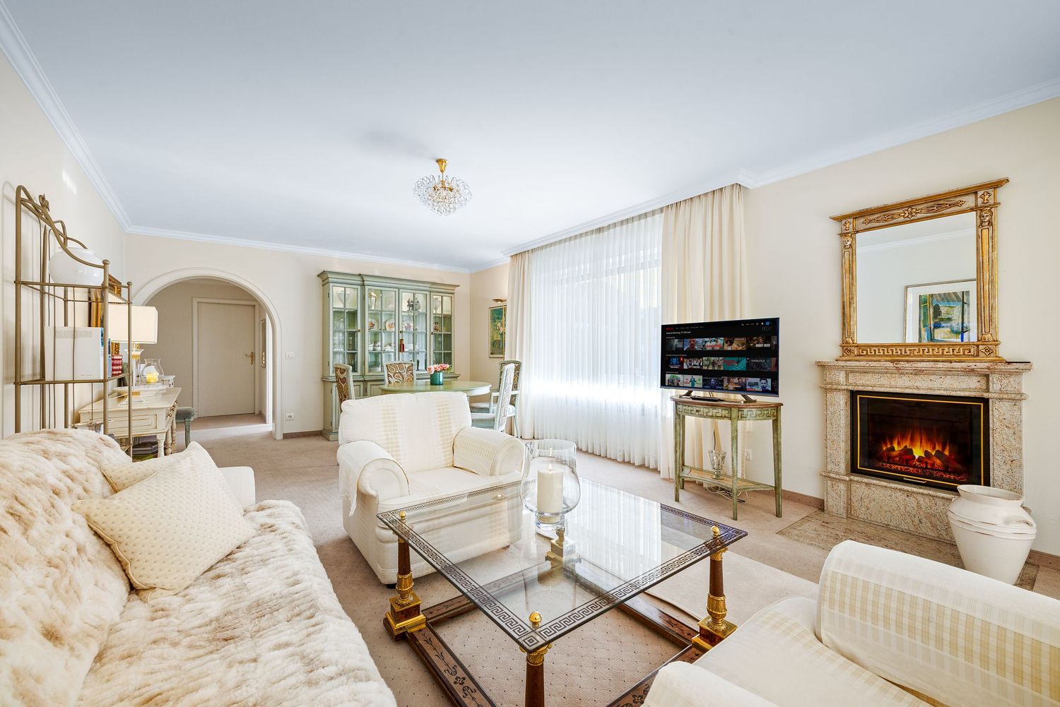 Luxurious white living room featuring an electric fireplace, ornate gold mirror, and cream colored furniture with glass tables.