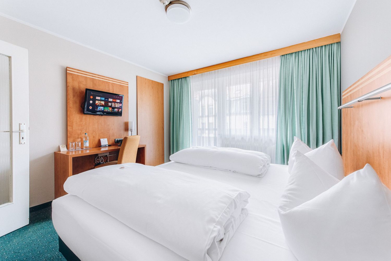Modern hotel room interior with white beds, wooden furniture, wall-mounted TV, and mint green curtains.
