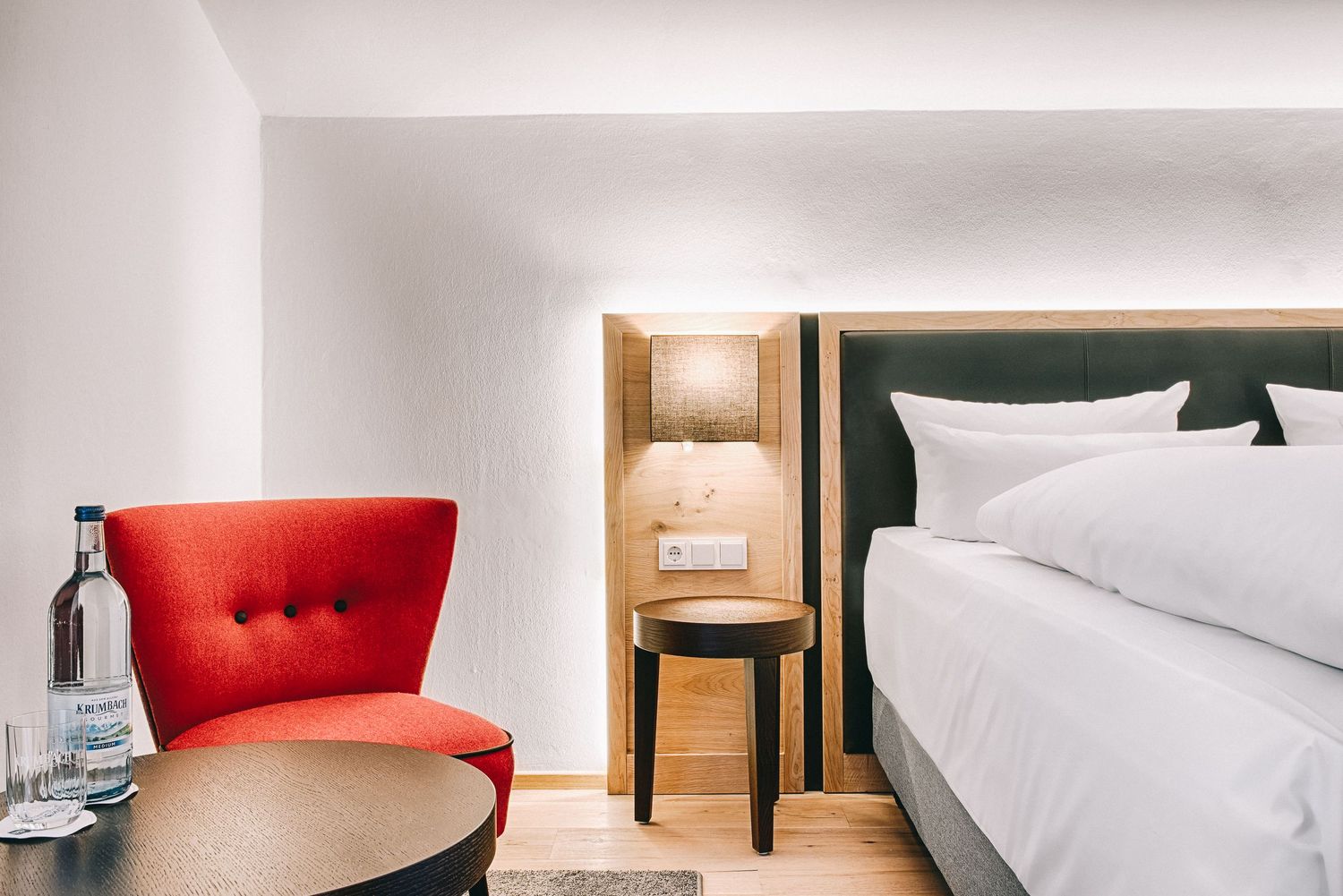 Modern hotel room interior with white bed, red accent chair, and warm wood bedside table.