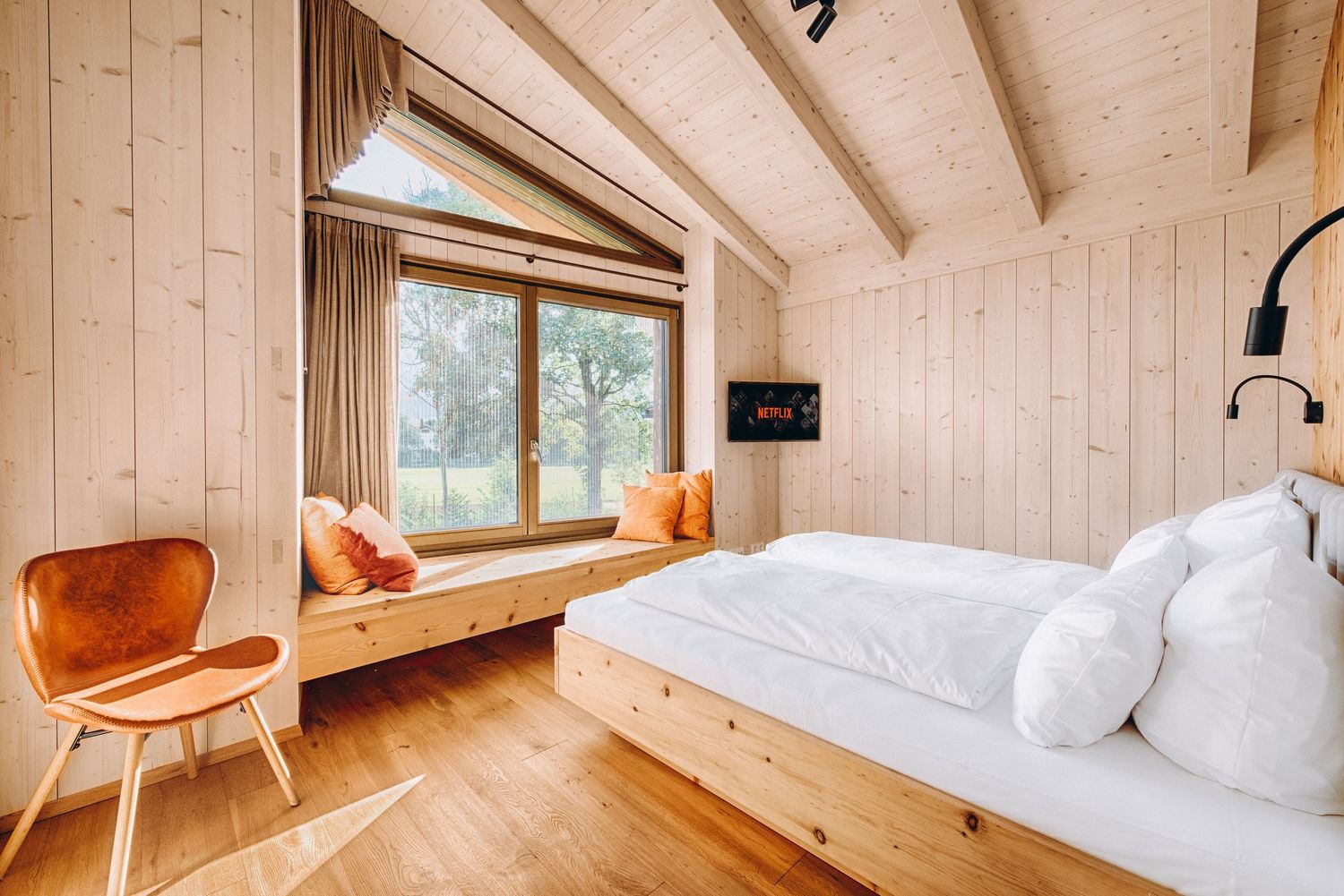 Modern wooden bedroom with slanted ceiling, built-in window seat, orange accent chairs and white bedding.