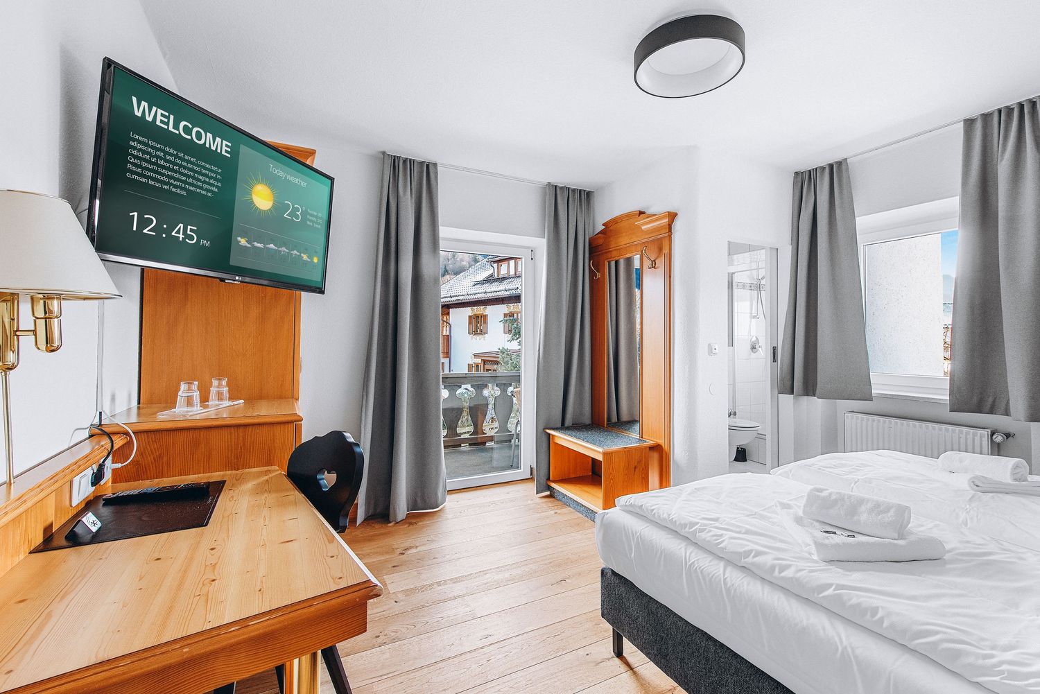 Modern hotel room with wooden desk, wall-mounted TV, double bed, and gray curtains in bright natural lighting.