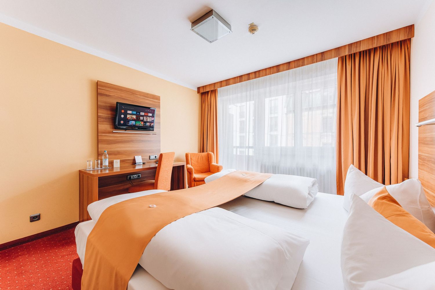 Modern hotel room with orange accents featuring a wall-mounted TV, white bed linens, and orange curtains.