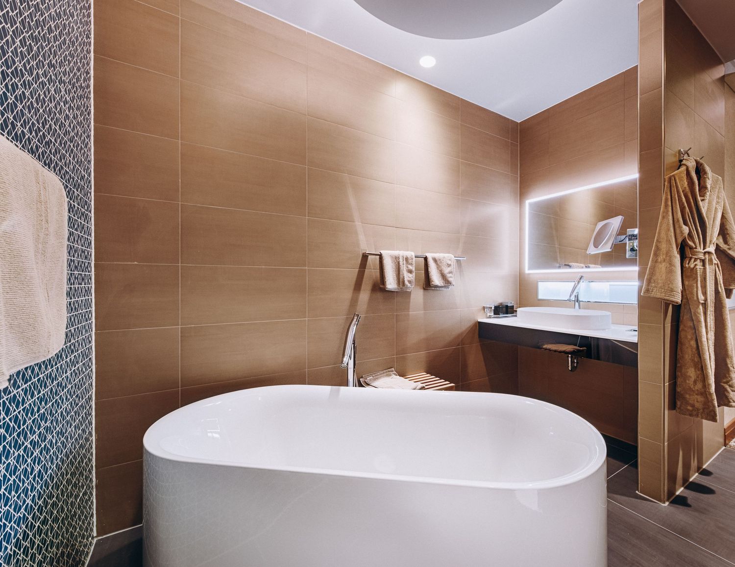 Modern luxury bathroom with oval freestanding tub, wood-paneled walls, and ambient lighting featuring plush bathrobes.
