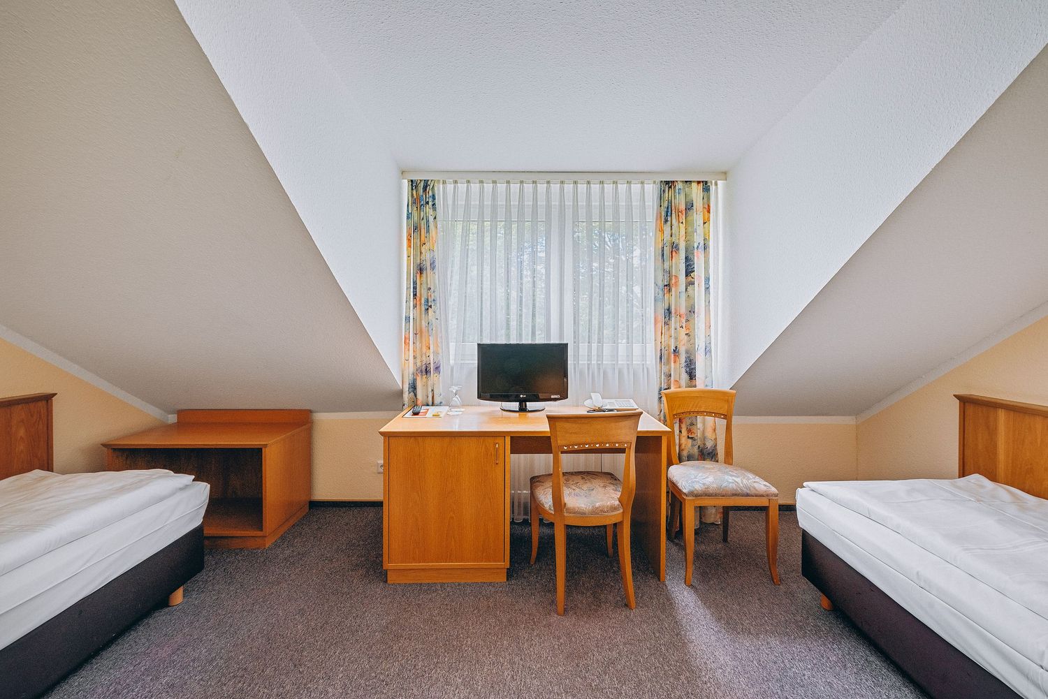 Basic hotel room with twin beds, wooden desk, and floral curtains under a sloped ceiling with white walls.