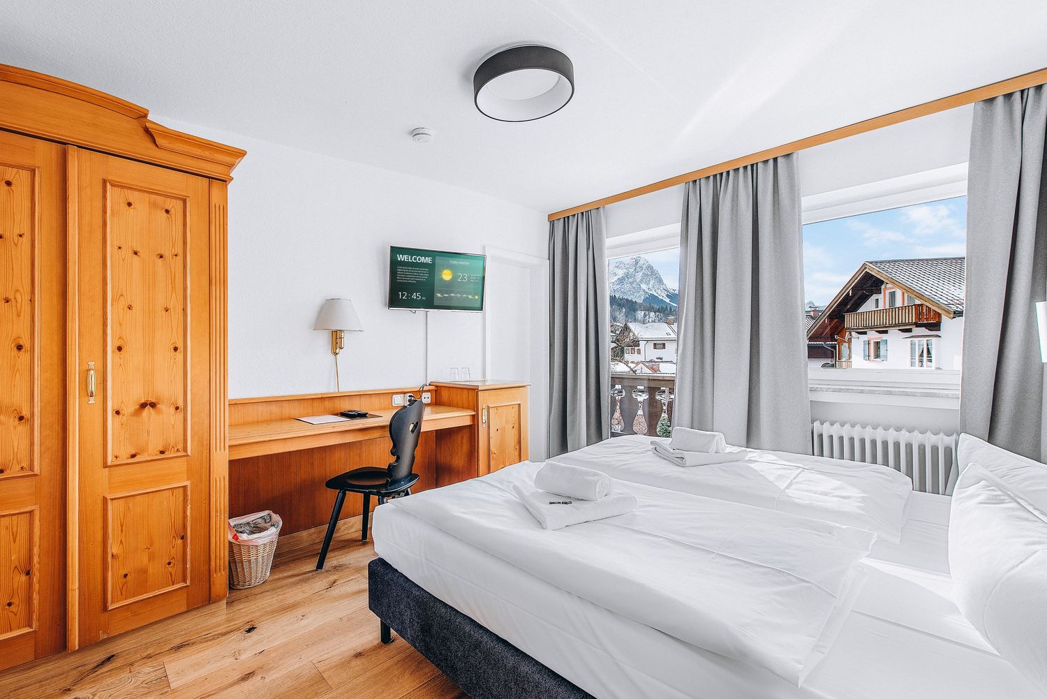 Modern mountain hotel room with wooden furnishings, white bedding, desk area and scenic window views of chalets.