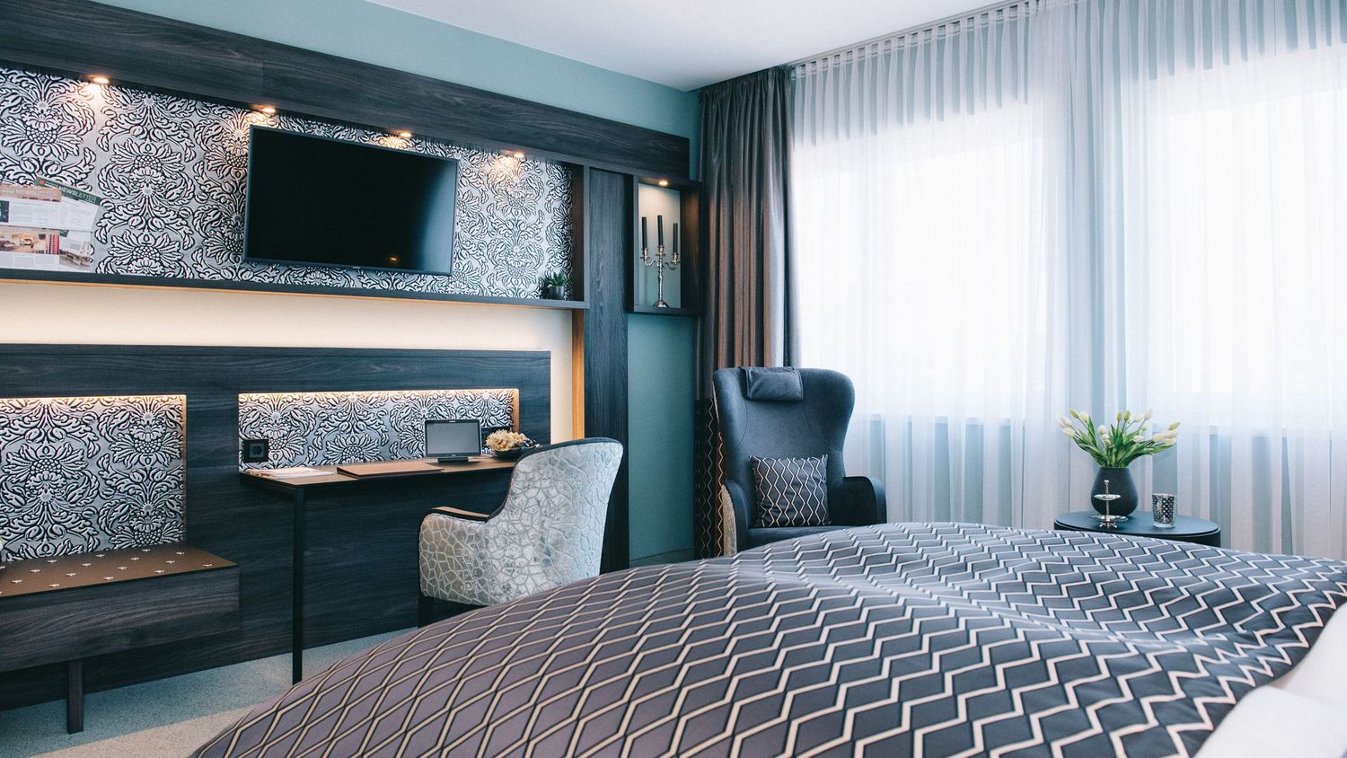 Modern hotel room with teal walls, a patterned accent wall, wall-mounted TV, and contemporary gray furnishings.