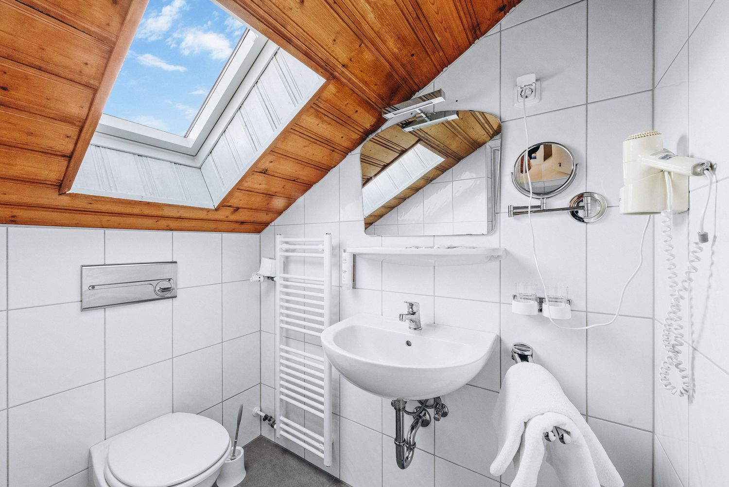 Attic bathroom with skylight, wooden ceiling panels, white tiles, and wall-mounted sink under sloped roof.