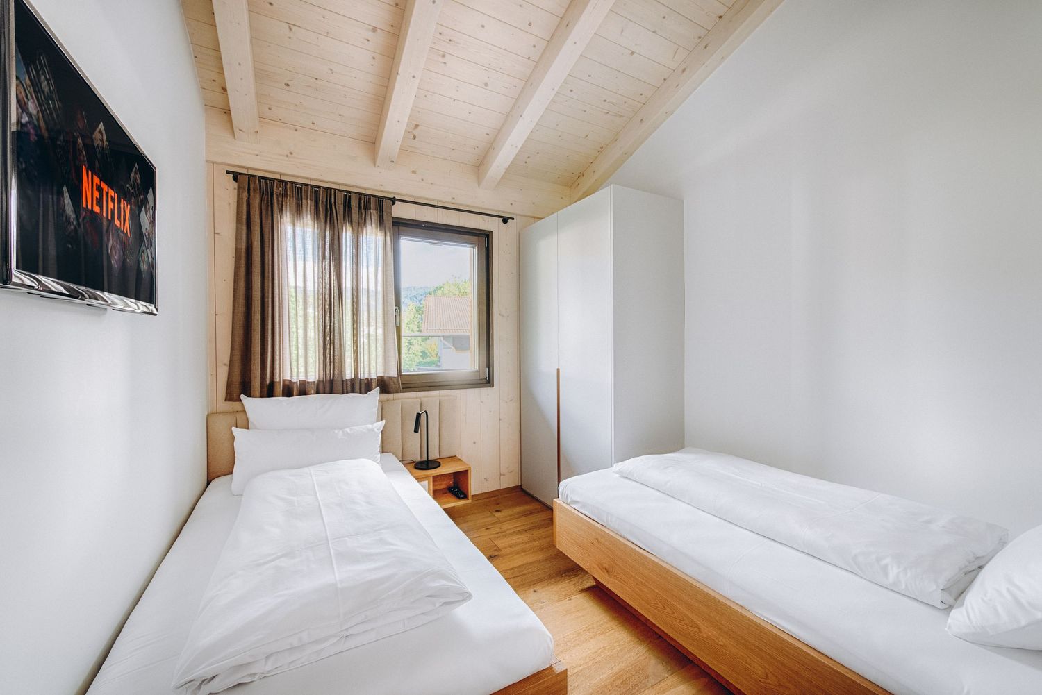 Modern minimalist bedroom with white walls, wooden floors, and natural light streaming through large windows.