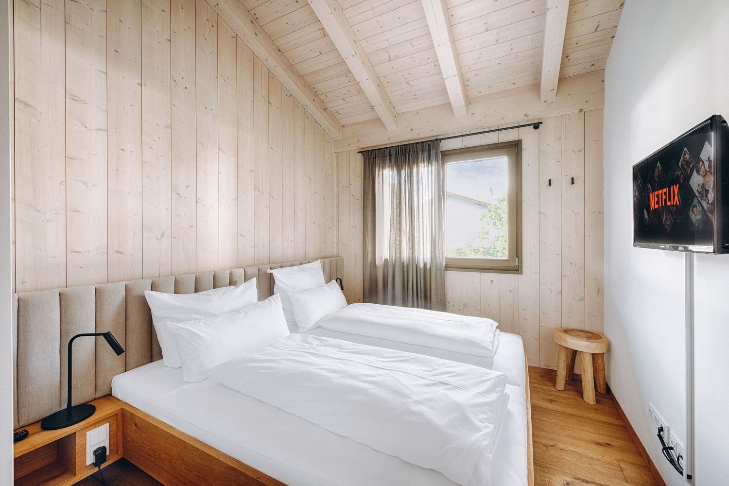 Modern wooden room with white paneled walls, exposed ceiling beams, crisp white bedding, and natural light.