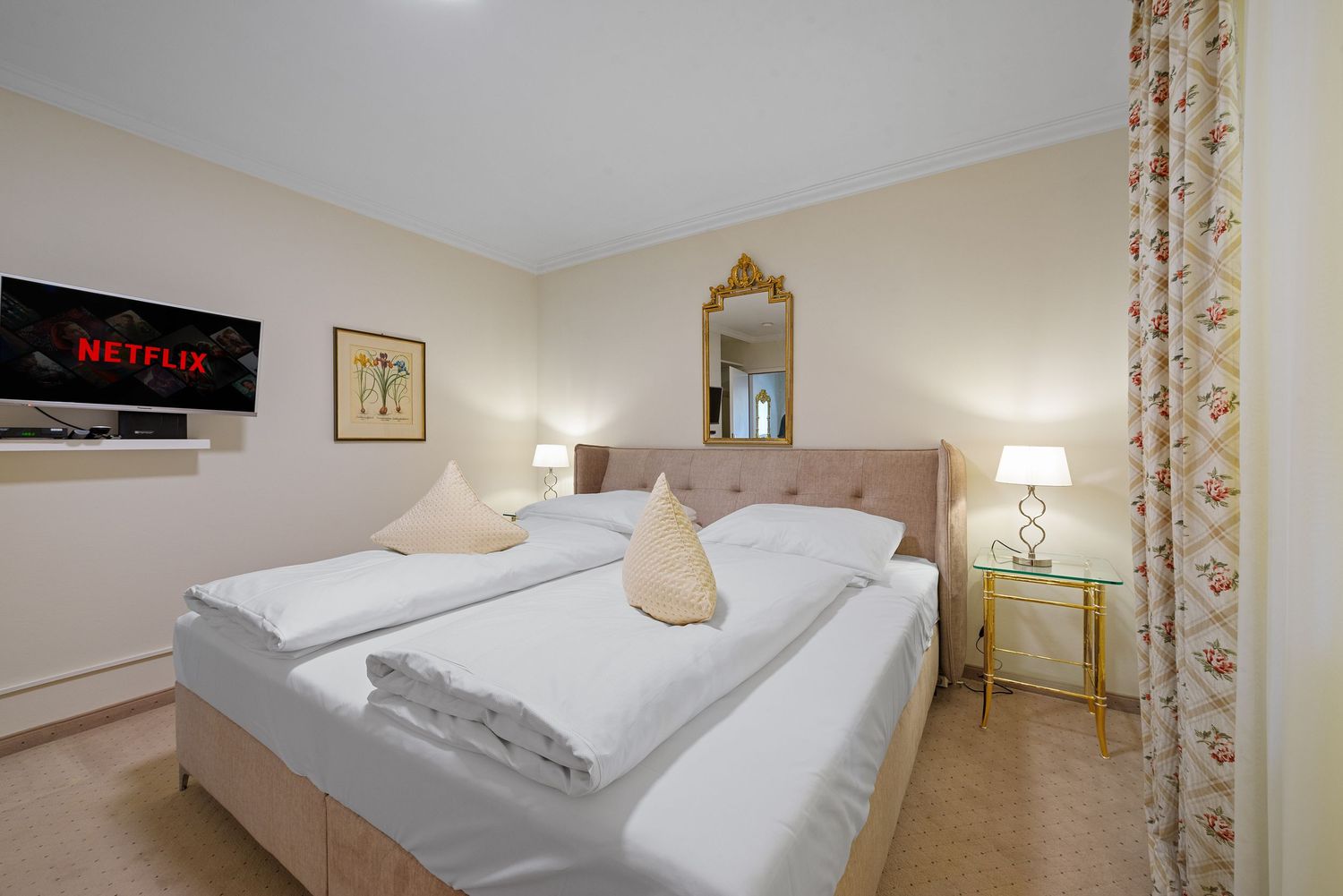 Modern hotel bedroom with beige walls, white bedding, decorative mirror, and wall-mounted display panel.