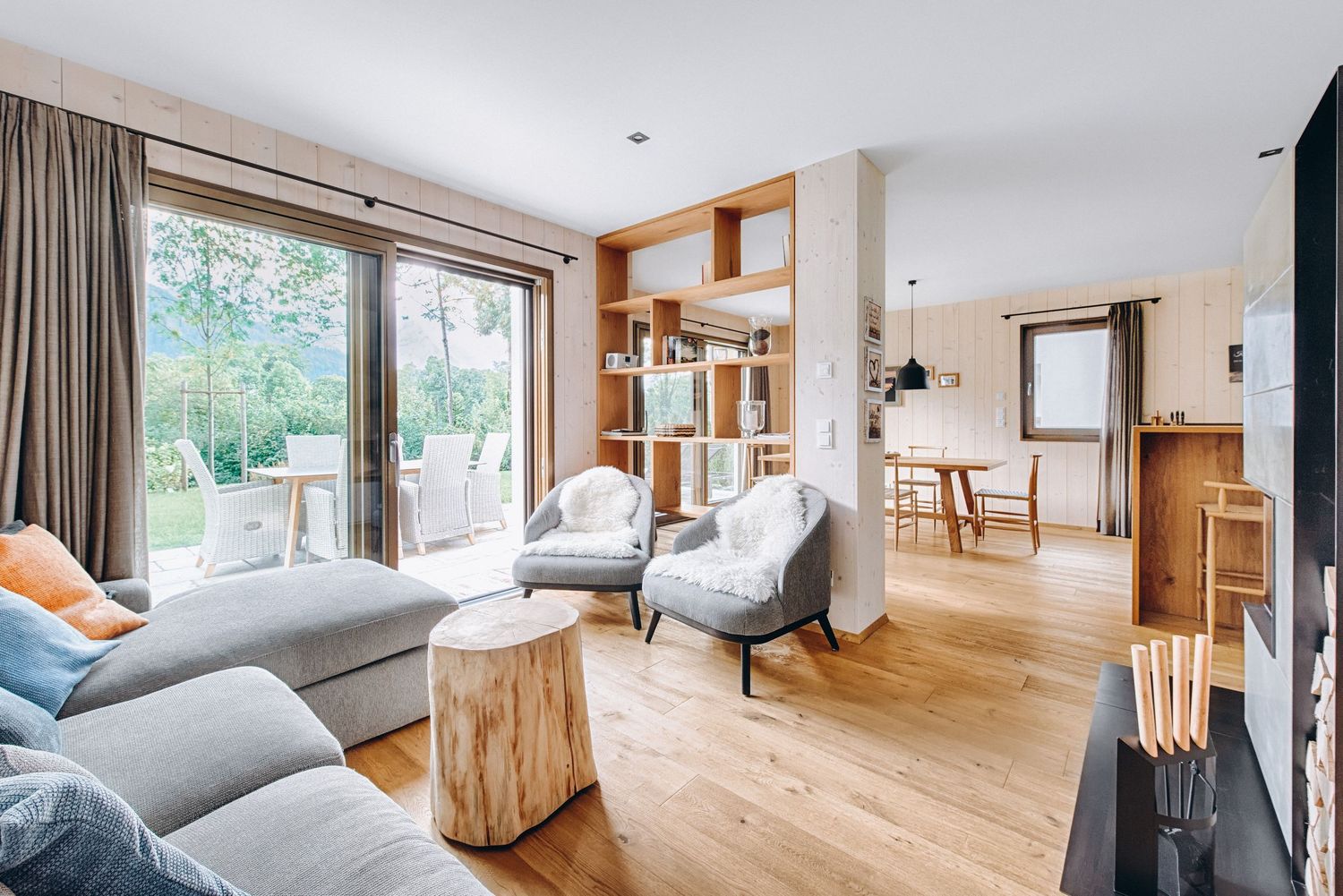 Modern living room interior with light hardwood floors, grey sectional sofa and floor-to-ceiling windows overlooking garden.