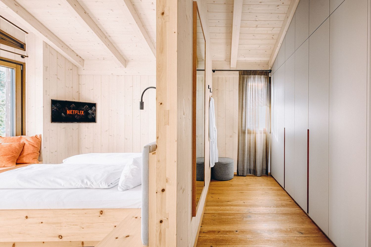 Modern bedroom interior with white walls, wooden floors, built-in wardrobes and orange accent pillows against white bedding.