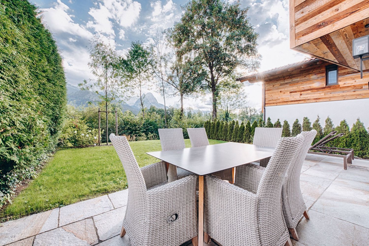 Modern outdoor patio furniture with wicker chairs and dining table on stone terrace overlooking landscaped garden.