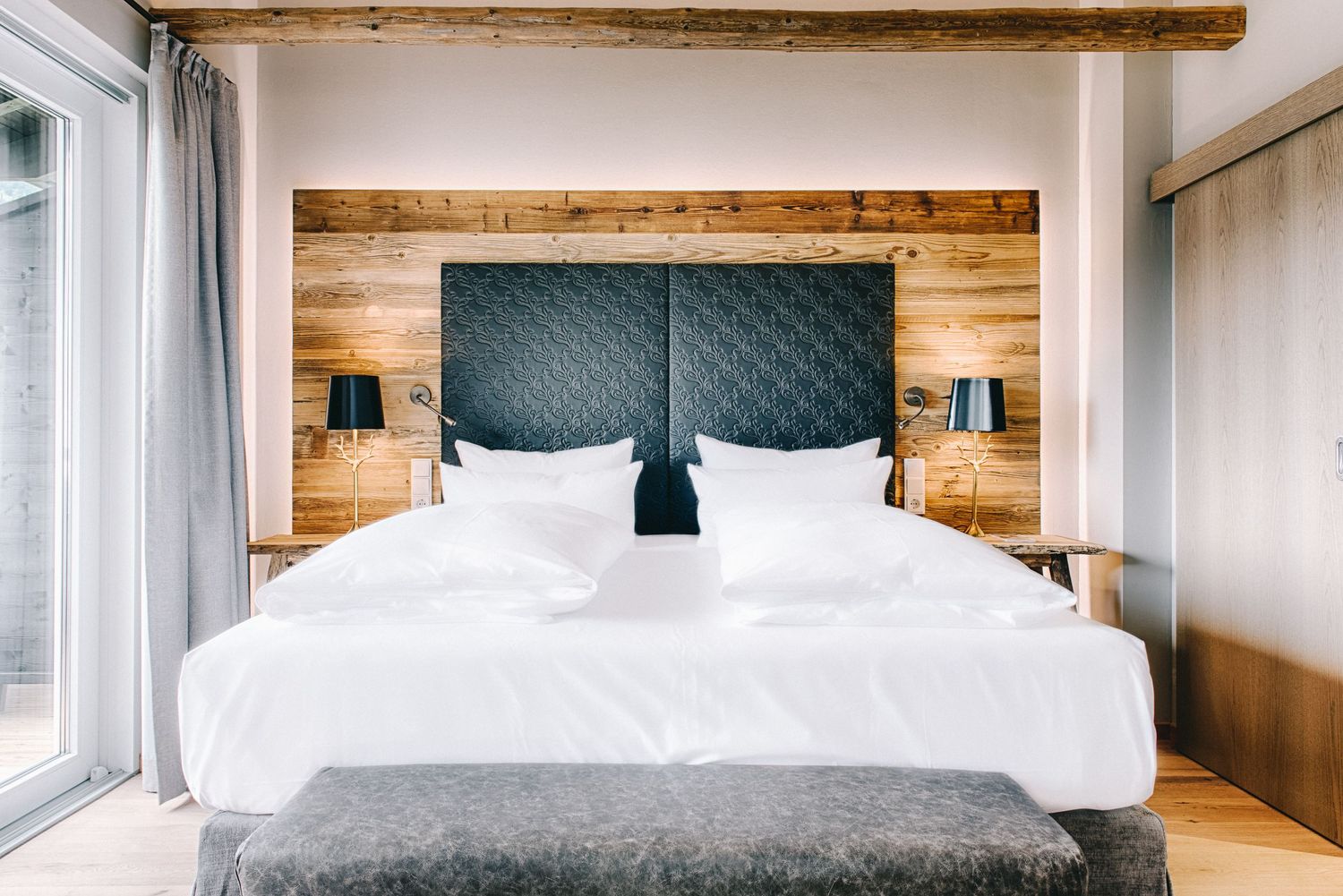 Modern hotel bedroom with wooden wall panels, tufted gray headboard, white bedding and minimalist decor.