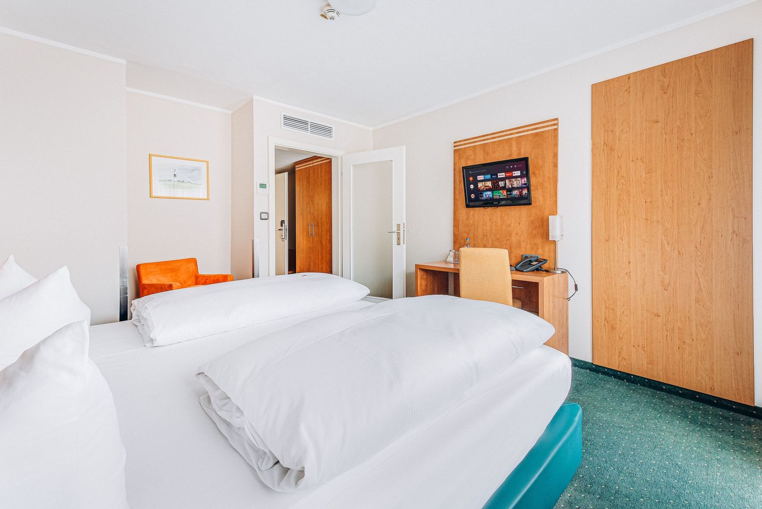 Contemporary hotel bedroom with teal bed base, wooden headboard, and orange accent wall.