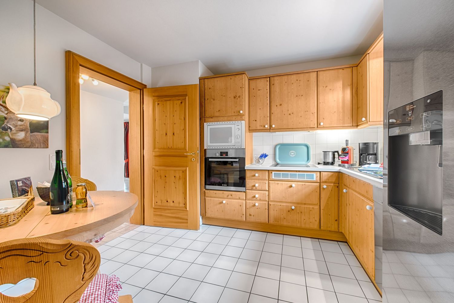 Modern kitchen interior with light wood cabinets, white tile flooring, and built-in appliances in an L-shaped layout.