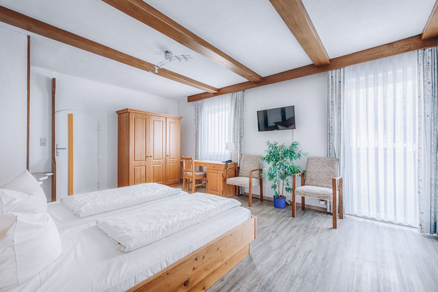 Bright hotel room with wooden beams, white bedding, hardwood floors, and wall-mounted TV near sheer curtained windows.