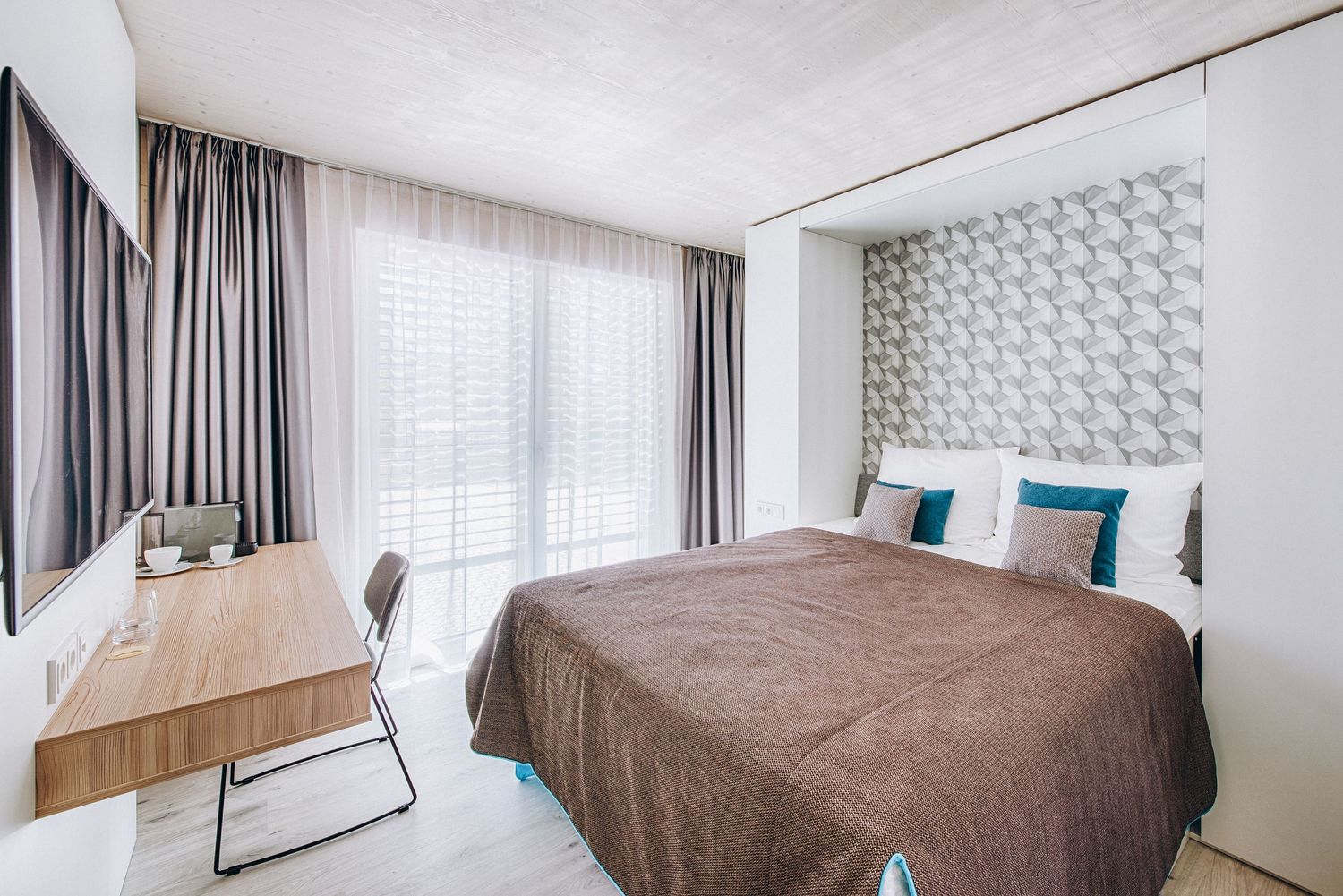 Modern minimalist hotel room with white walls, brown bed, wooden desk and geometric headboard pattern.
