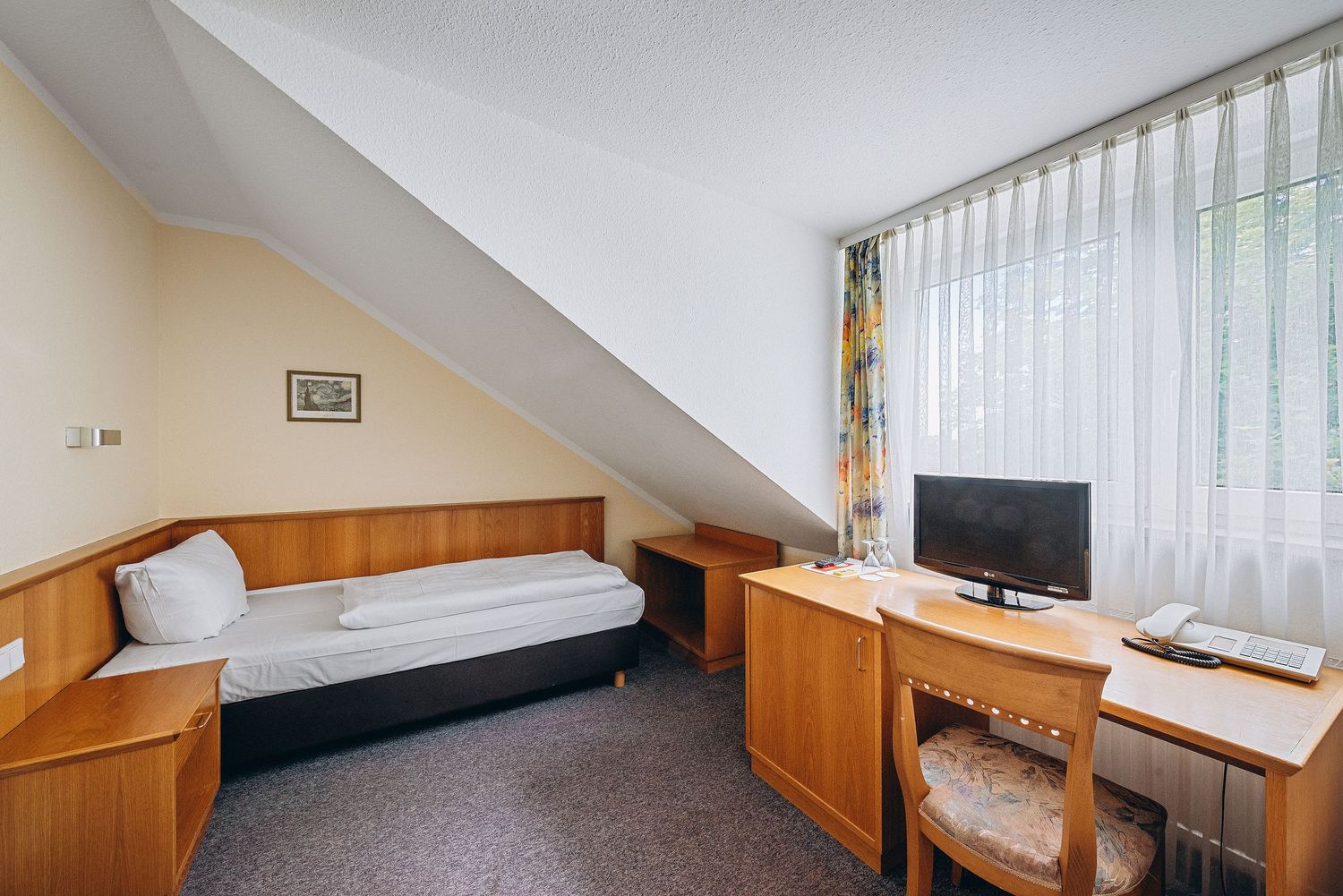 Modern hotel room interior with sloped ceiling featuring a single bed, wooden desk, and TV under natural lighting.