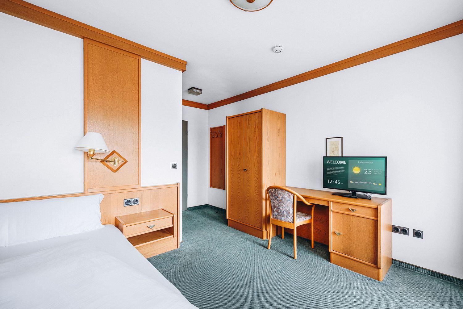 Modern hotel room with wooden furniture, green carpet, white walls and a flat-screen TV mounted on the wall.