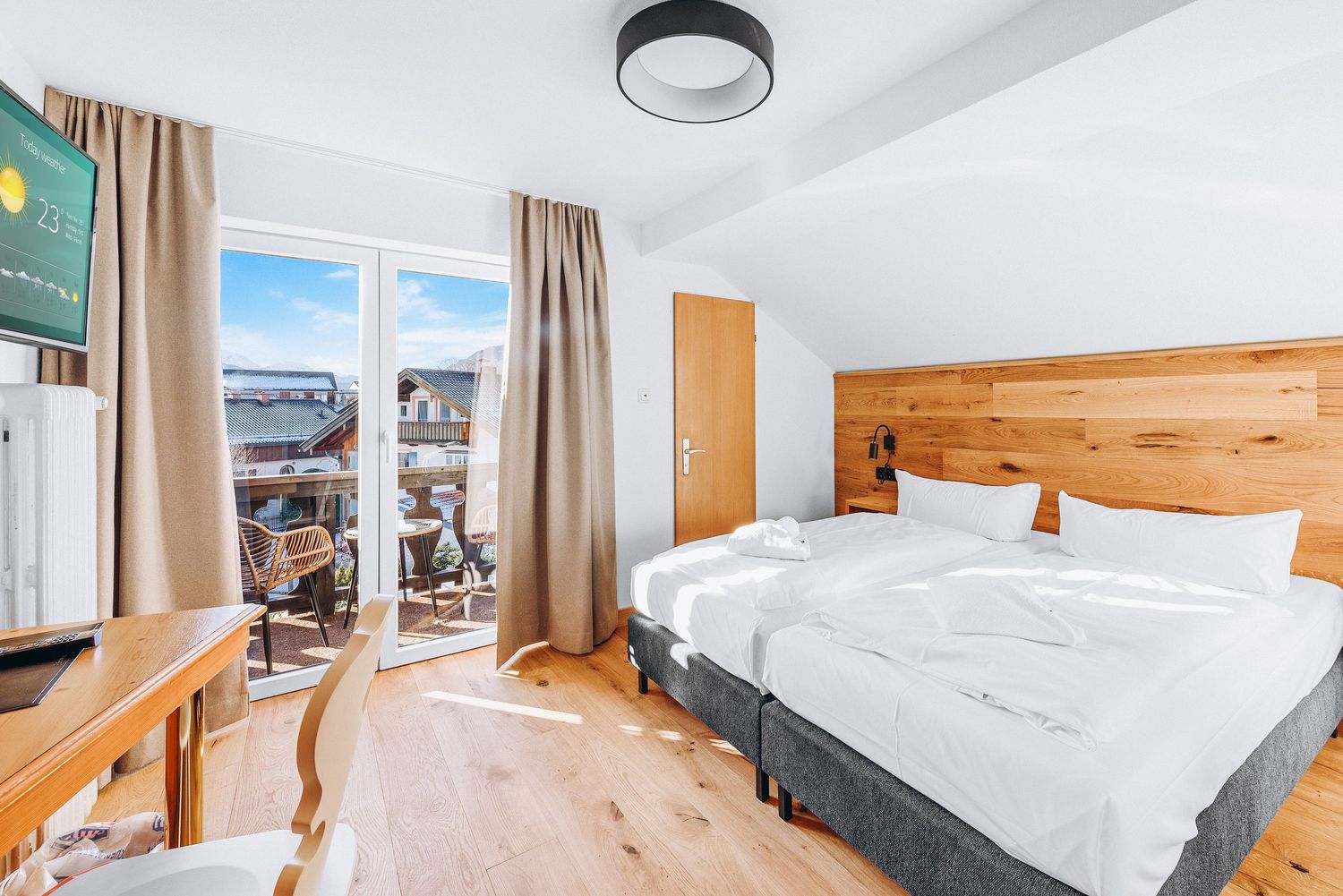 Modern hotel room with wooden headboard, twin beds, beige curtains, mounted TV and stunning city view through large window.