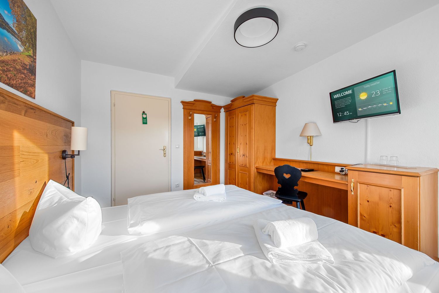 Bright hotel bedroom with wooden headboard, white bedding, built-in desk, and mounted television.
