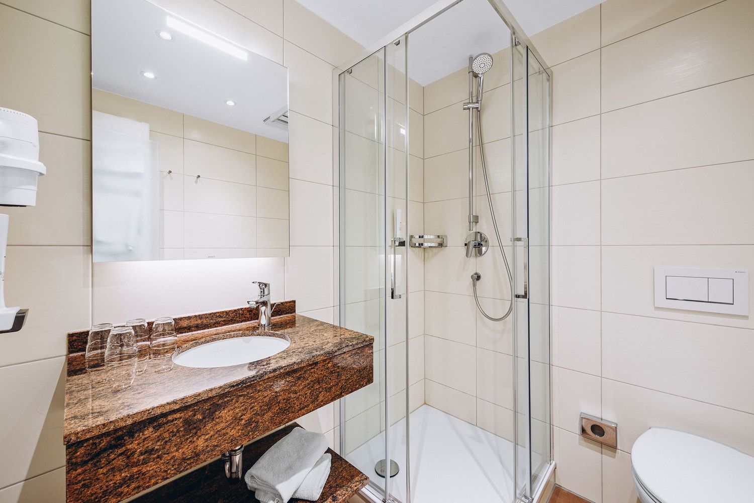 Modern hotel bathroom with glass shower enclosure, brown marble vanity counter, and beige tiled walls.