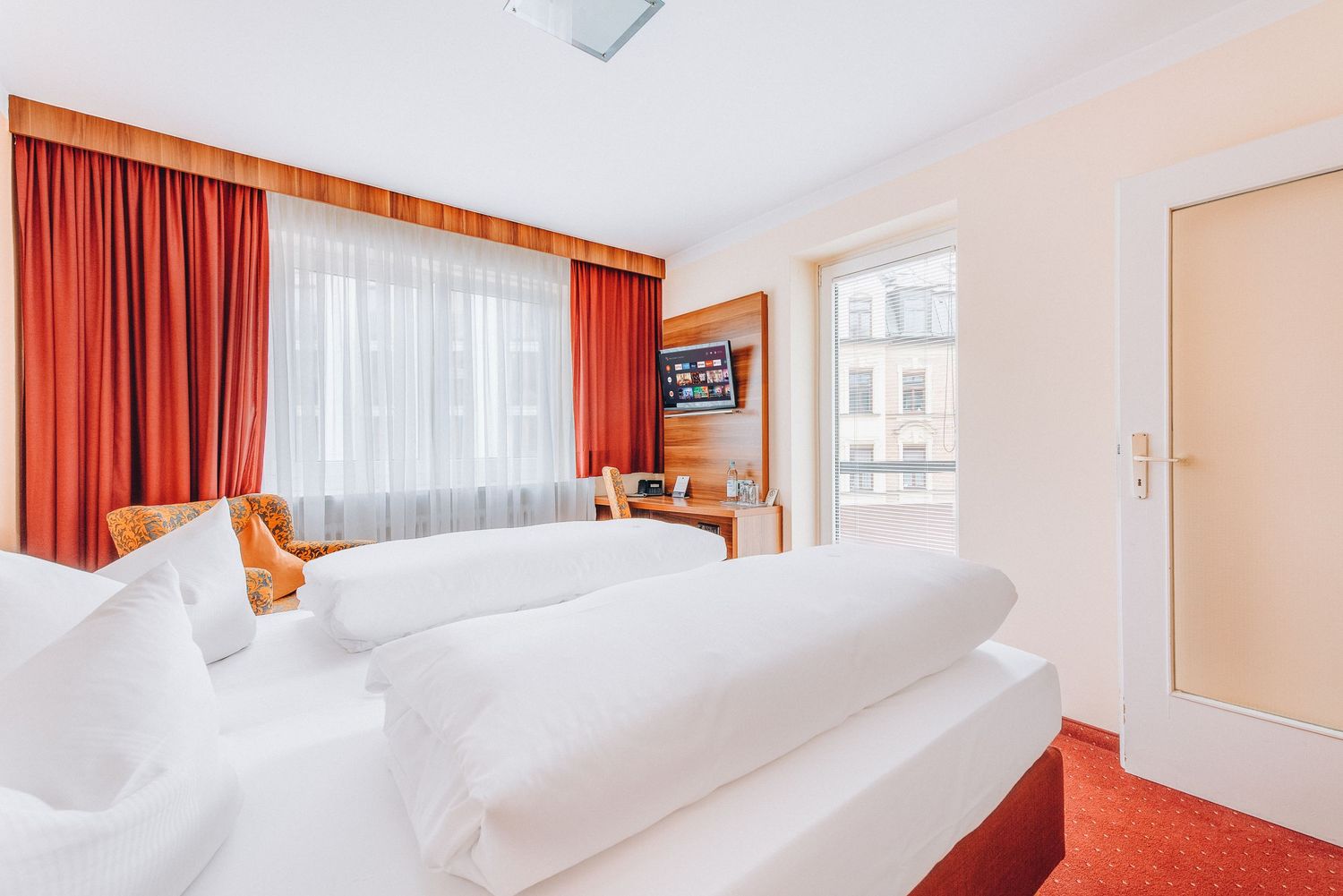 Bright hotel room with red curtains, white bedding, and large windows offering city views.