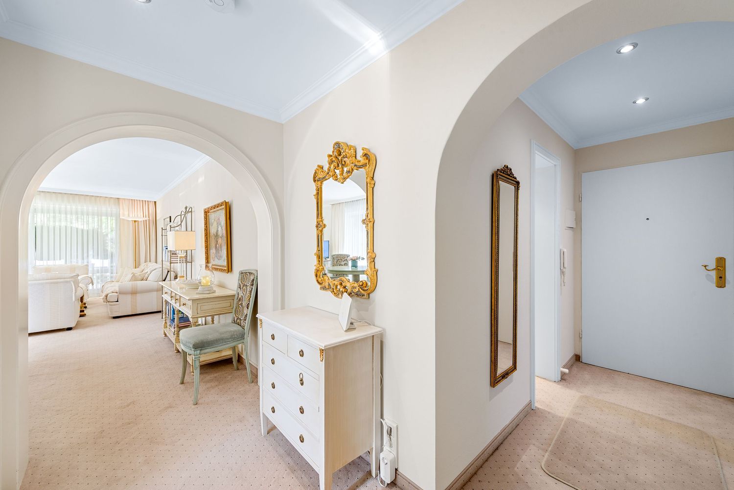 Elegant hallway with arched doorway features ornate gold mirror, white chest of drawers, and beige carpet.