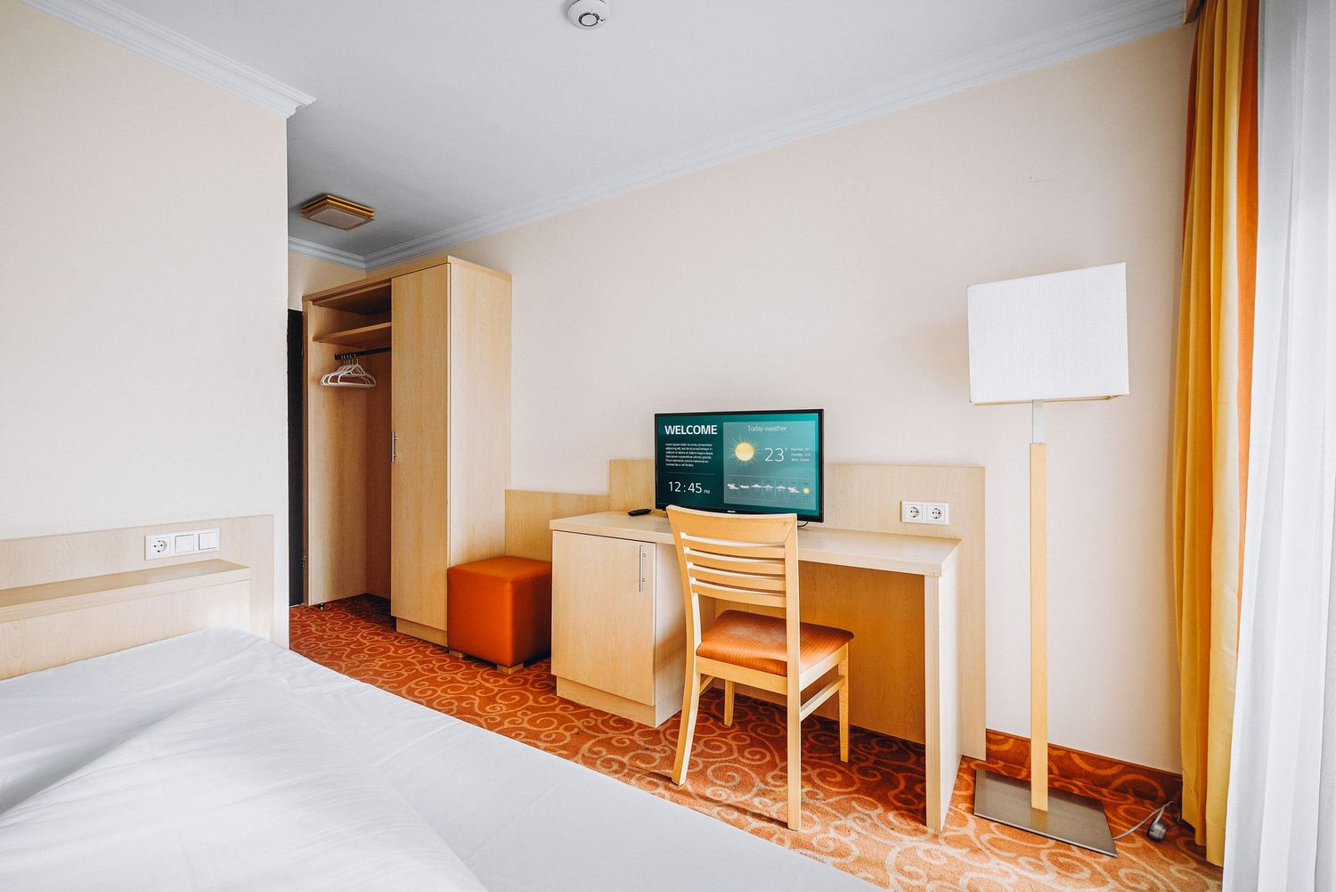 Modern hotel room interior with single bed, desk area, flat screen TV, and orange carpeting illuminated by natural light.