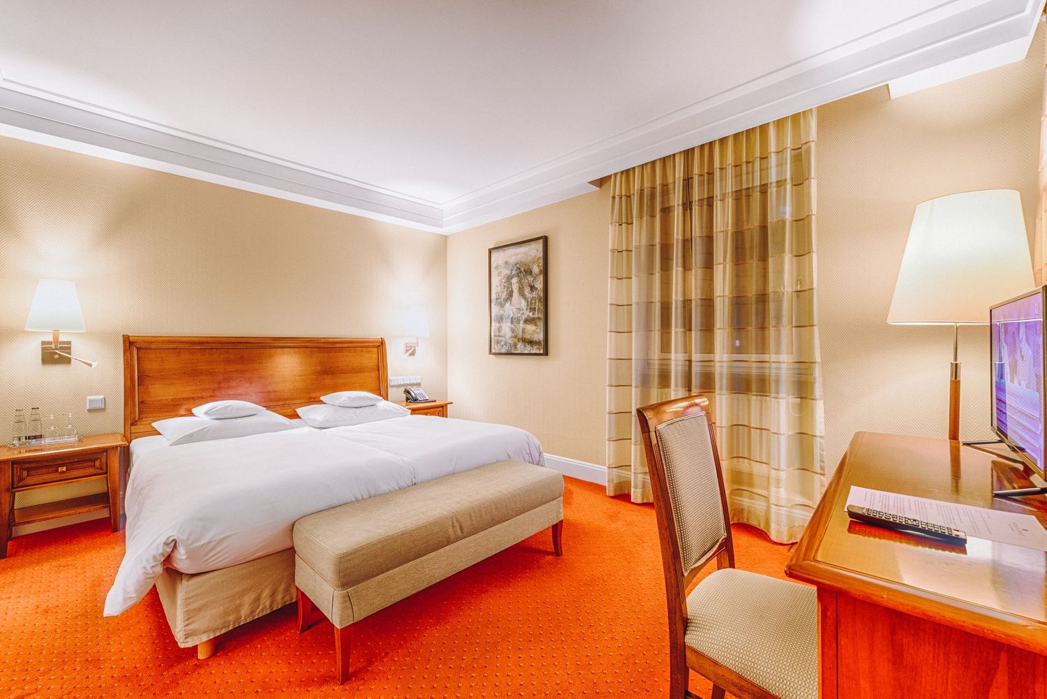 Contemporary hotel room featuring wooden headboard, red carpet, and sheer curtains with warm lighting.