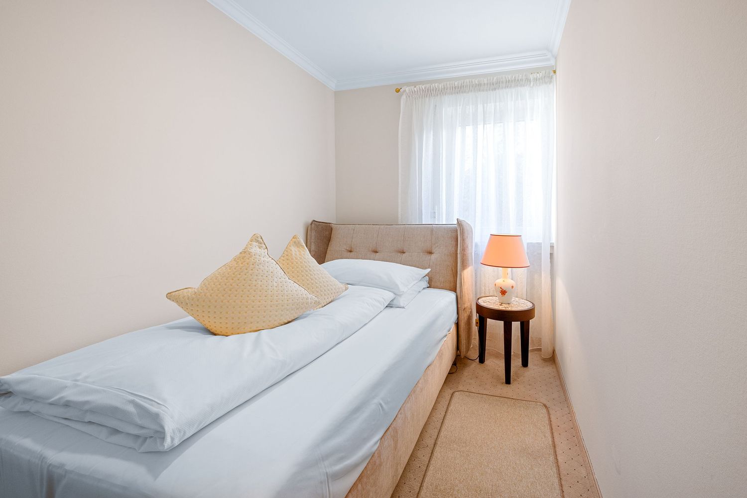 Minimalist hotel bedroom with tan walls features a neatly made single bed, accent chair, and orange table lamp.