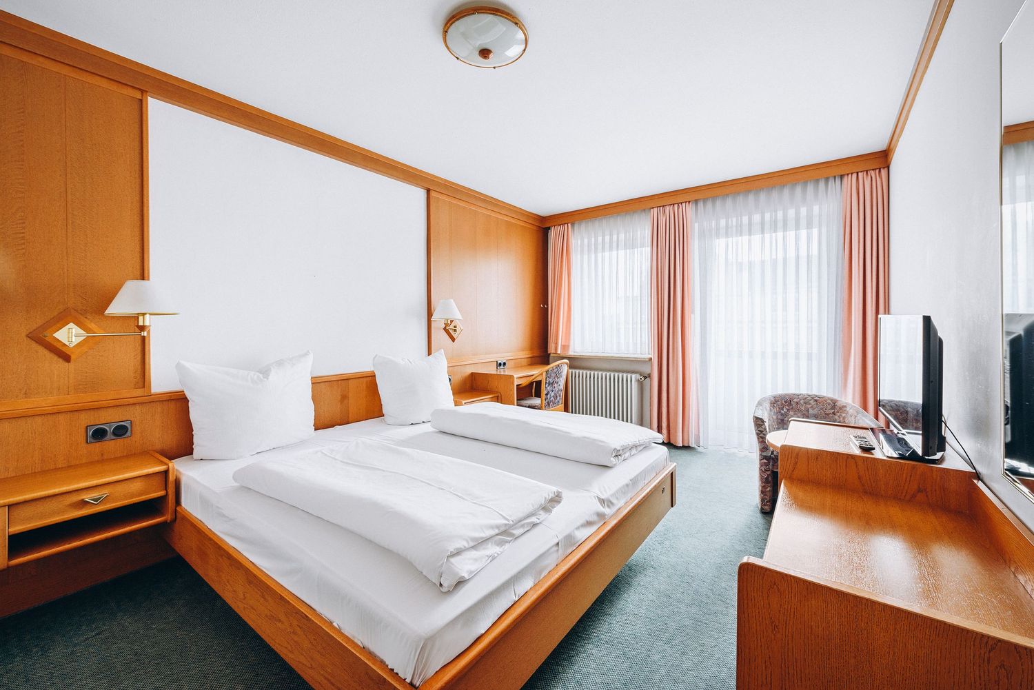 Bright hotel room interior with wooden furniture, white bedding, green carpet and peach curtains near windows.