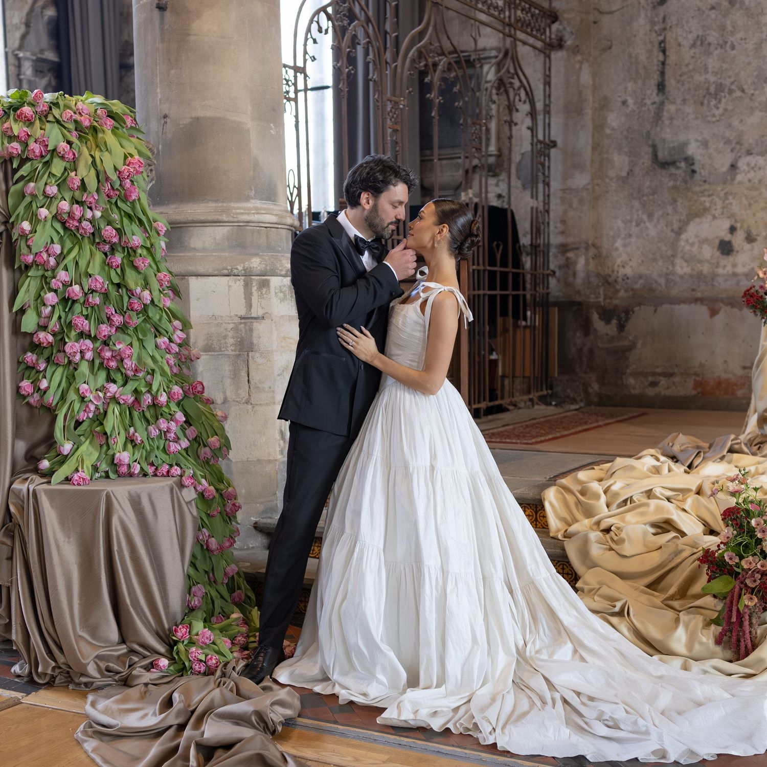 A romantic wedding couple shares a tender kiss in a rustic stone venue decorated with pink floral arrangements.