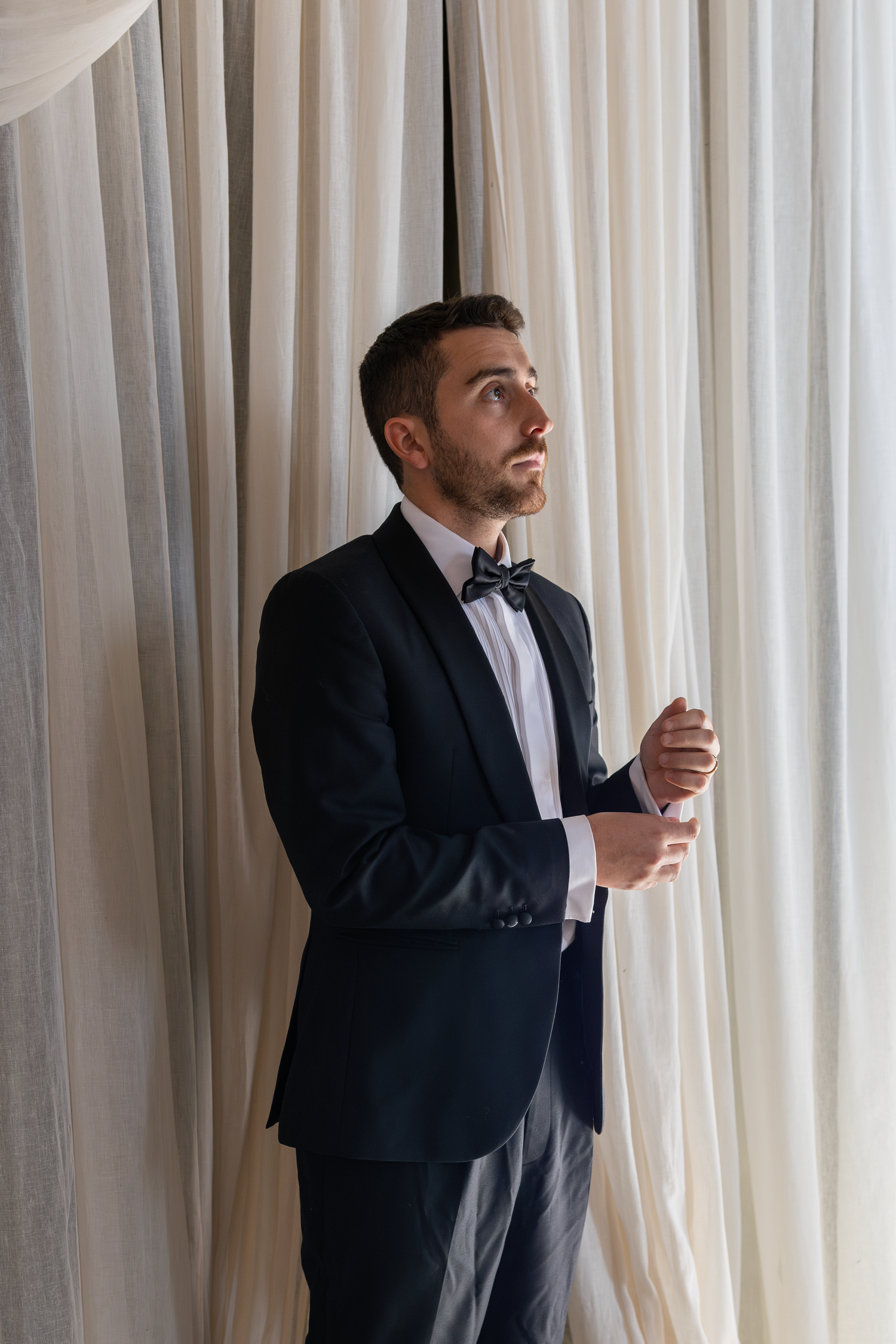 A groom in a black tuxedo adjusts their cuffs by white curtains.
