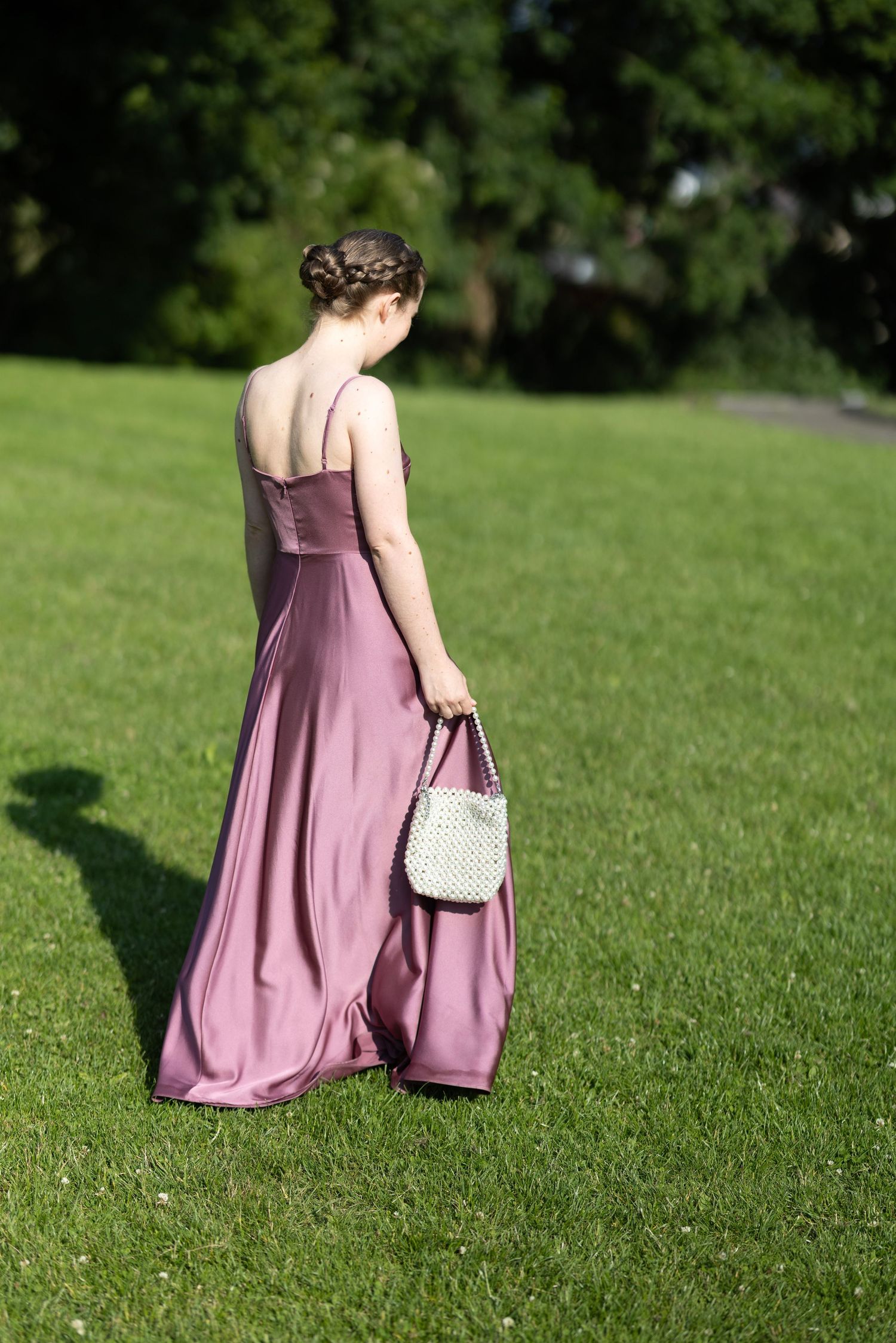 Person in a flowing mauve dress walking on lush green grass with a white pearl handbag.