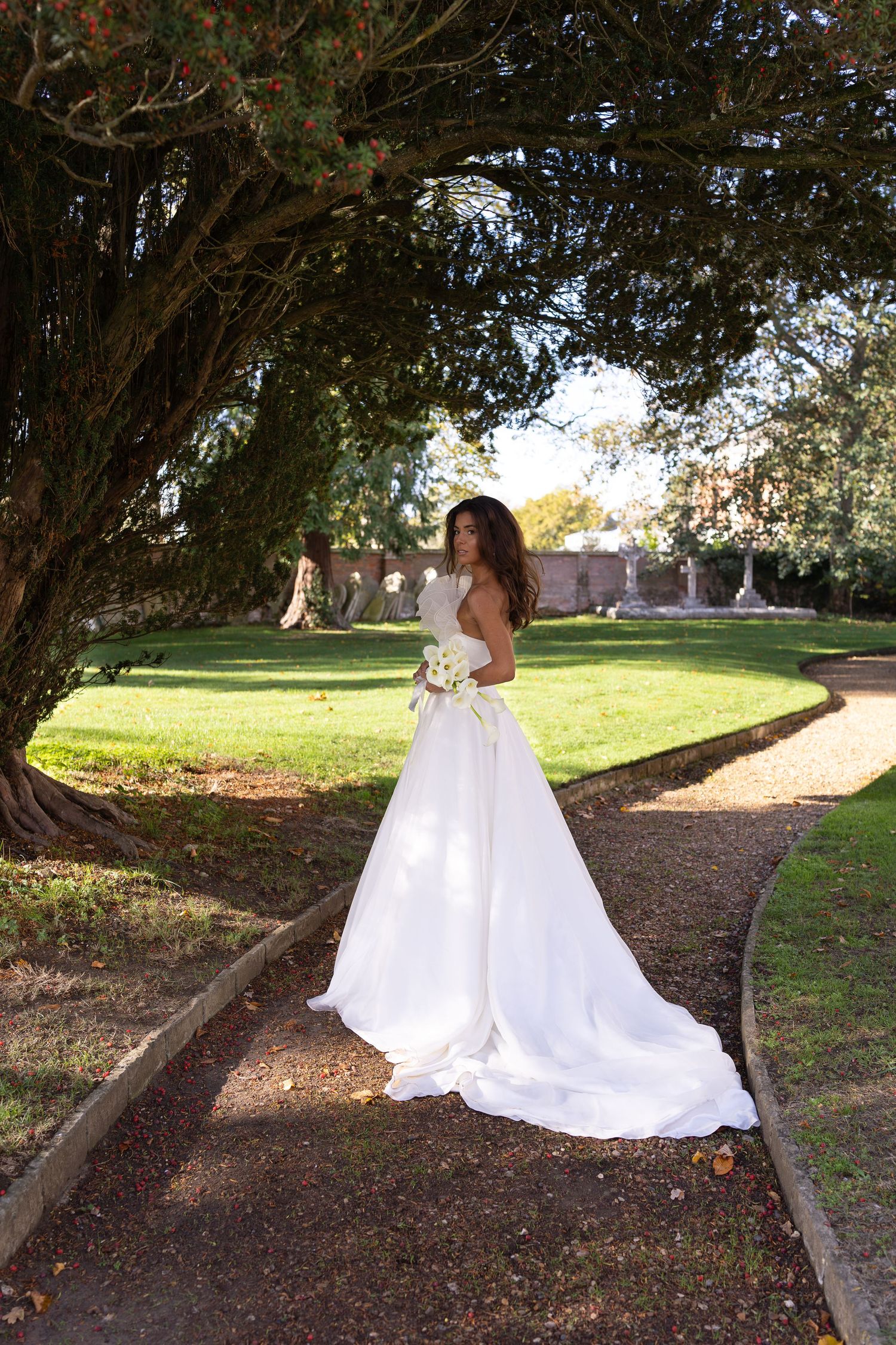 A bride in a white wedding dress poses on a garden path under overhanging trees.