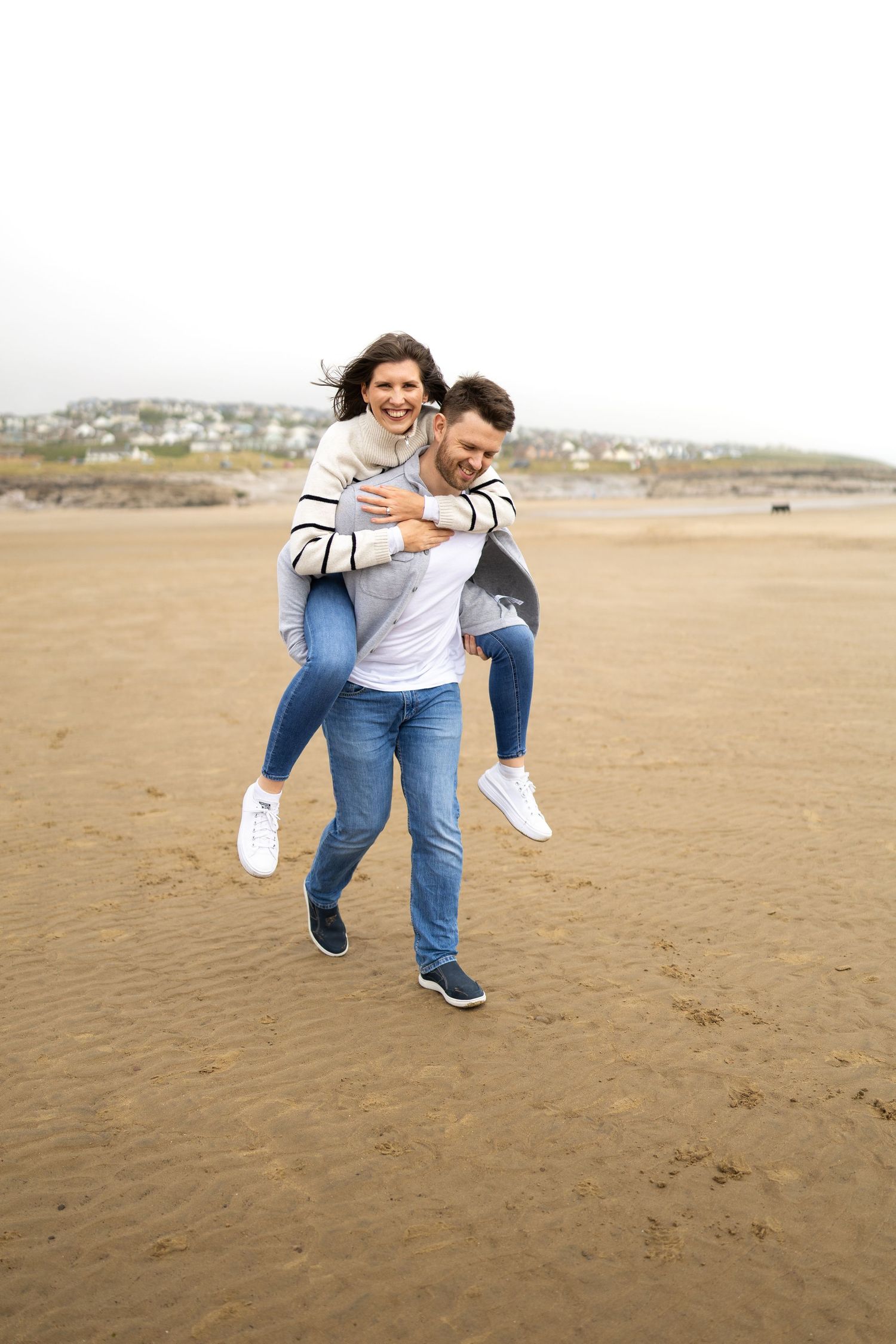 Playful couple running and laughing together on a sandy beach during a casual photoshoot.
