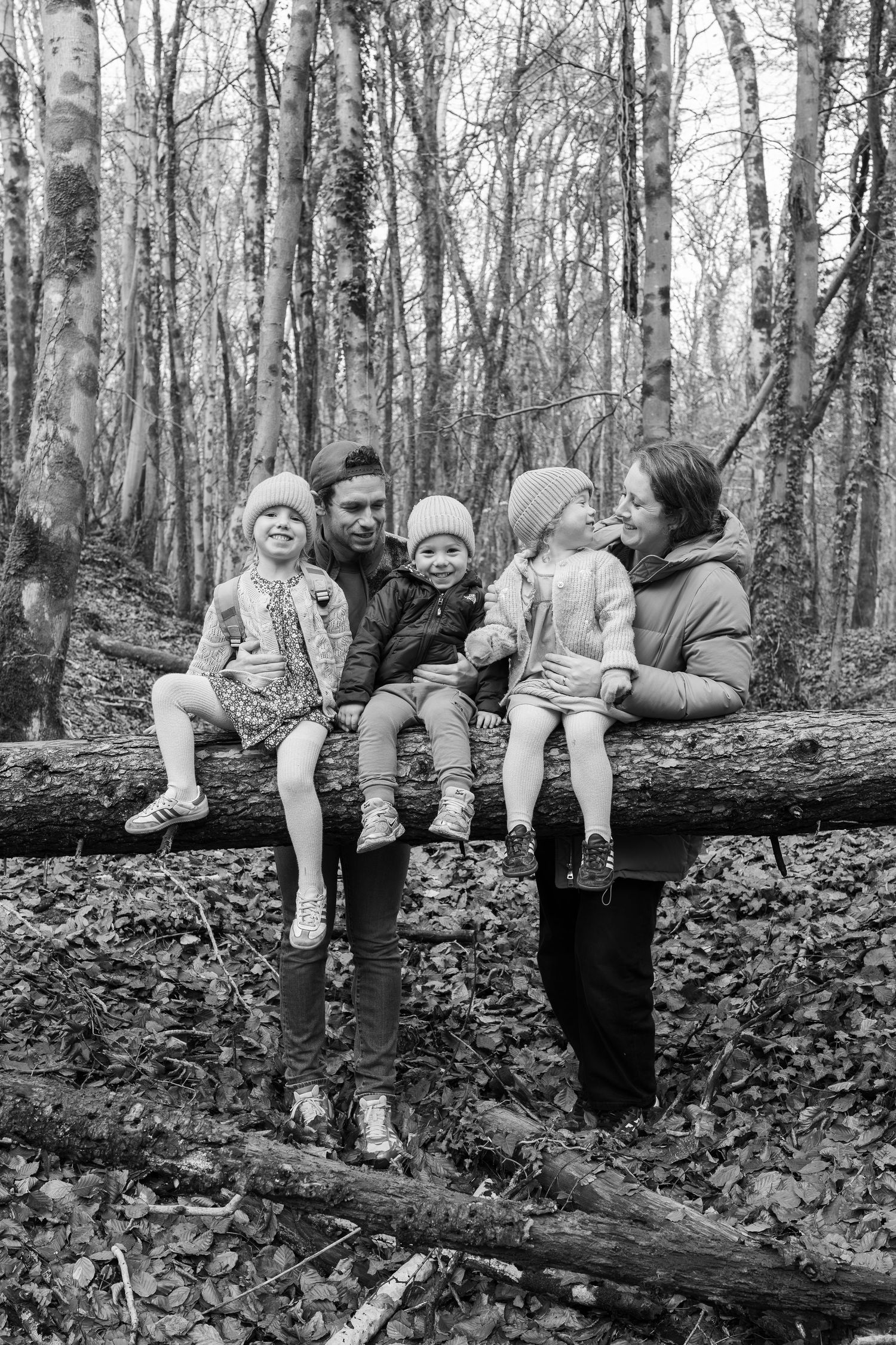 People sit casually on a large fallen tree trunk in a leafy forest setting.