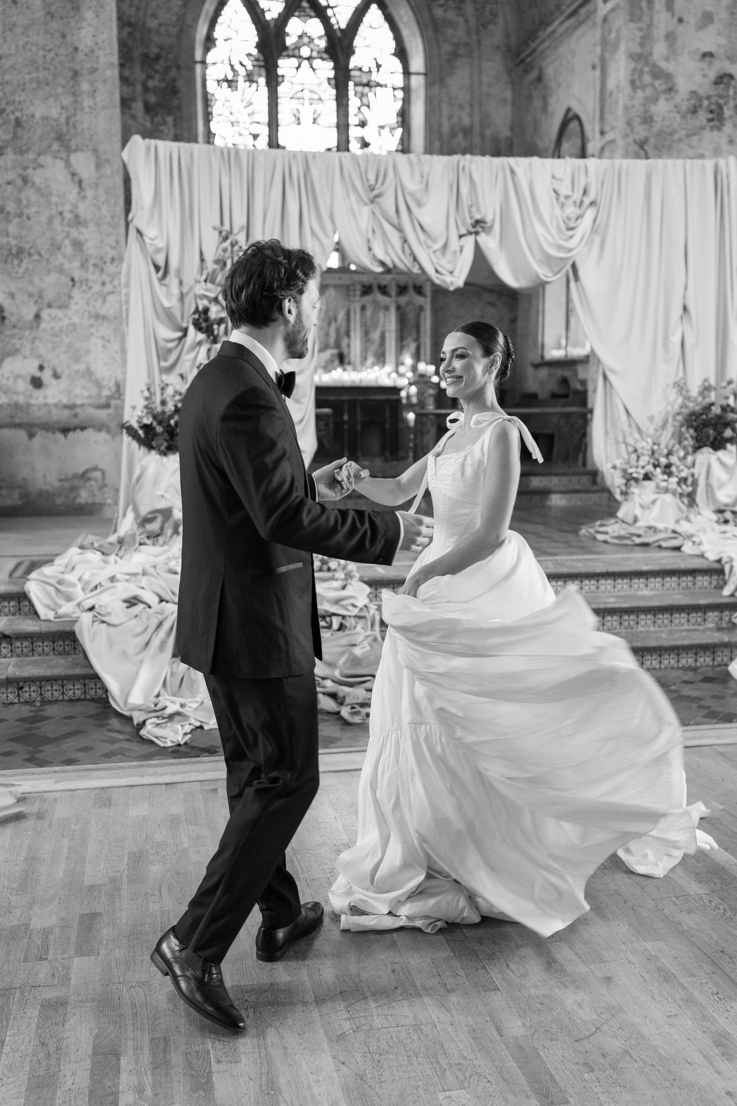 A couple shares their first dance in a historic church venue with gothic windows and flowing white drapery decorations.