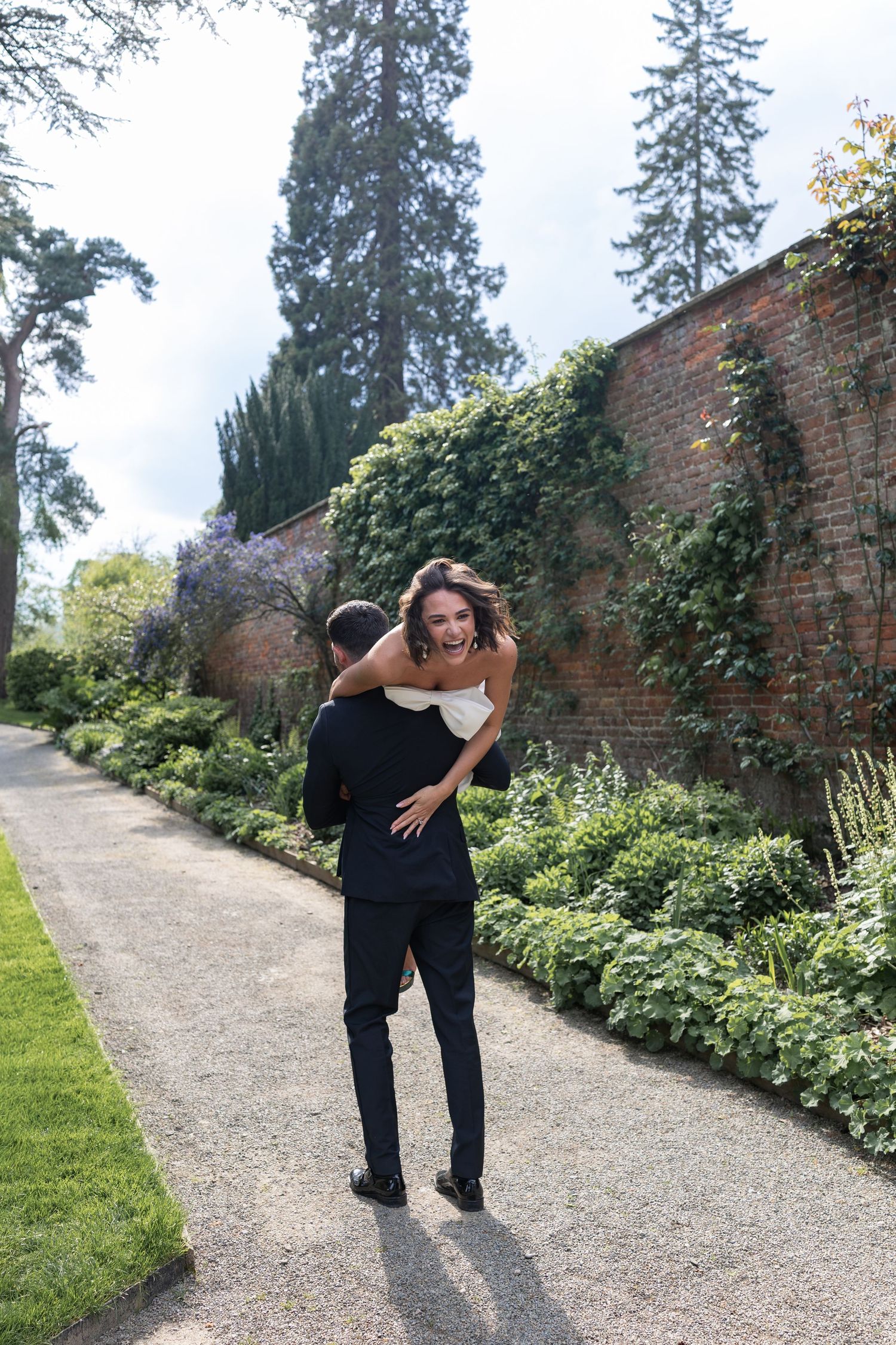 Couple laughing and embracing on a garden path surrounded by ivy-covered brick walls and lush greenery.