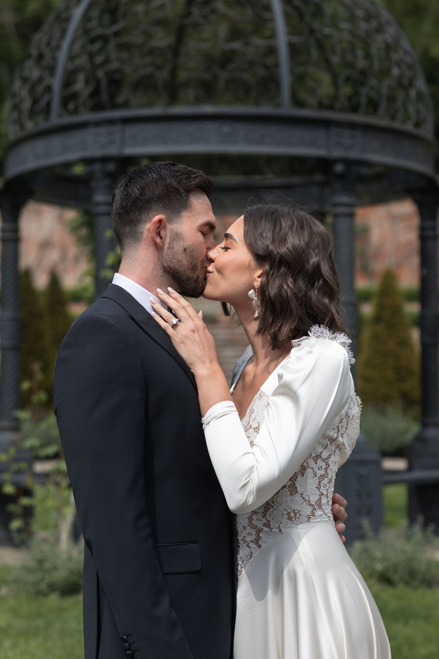 A bride and groom share a romantic kiss outdoors near an ornate gazebo on their wedding day.