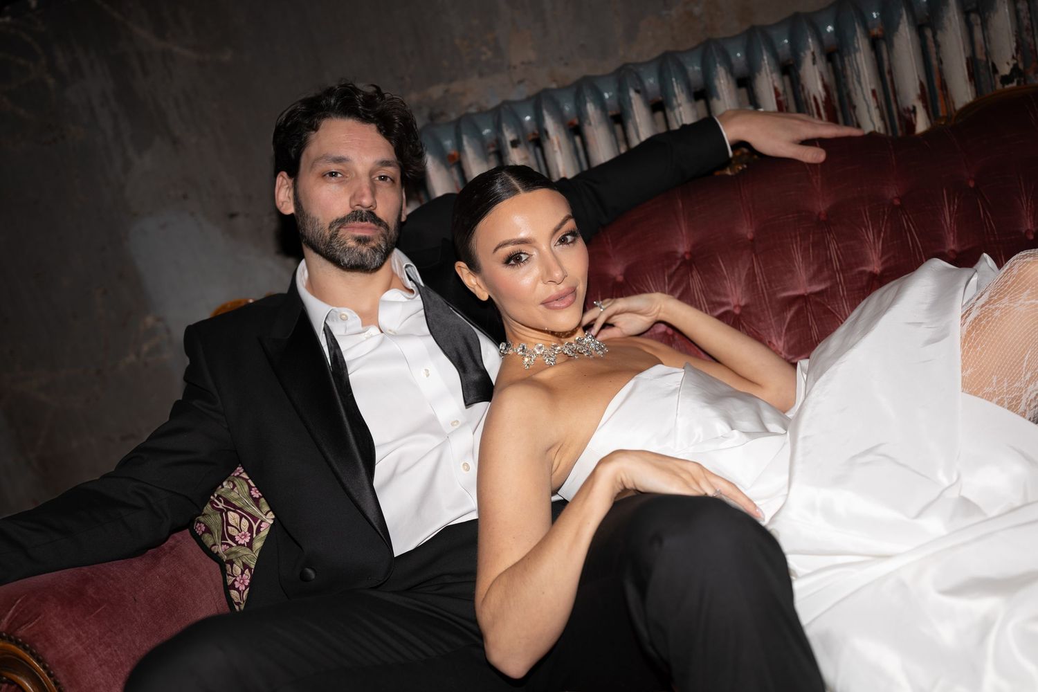 A couple in formal attire poses together on a red leather couch at an elegant event.