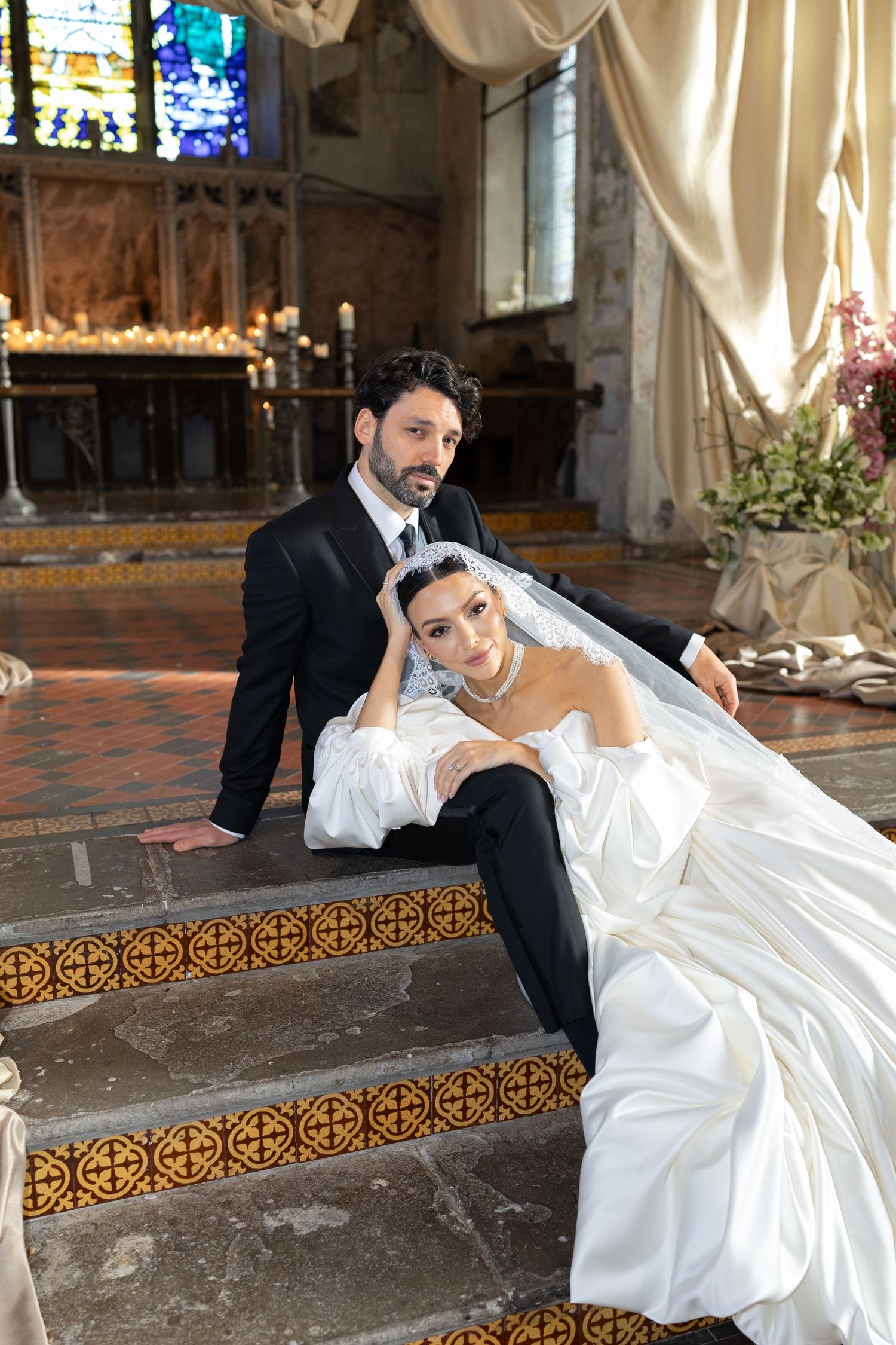 A bride in white gown reclines elegantly on ornate church steps while the groom sits nearby in a dark suit.