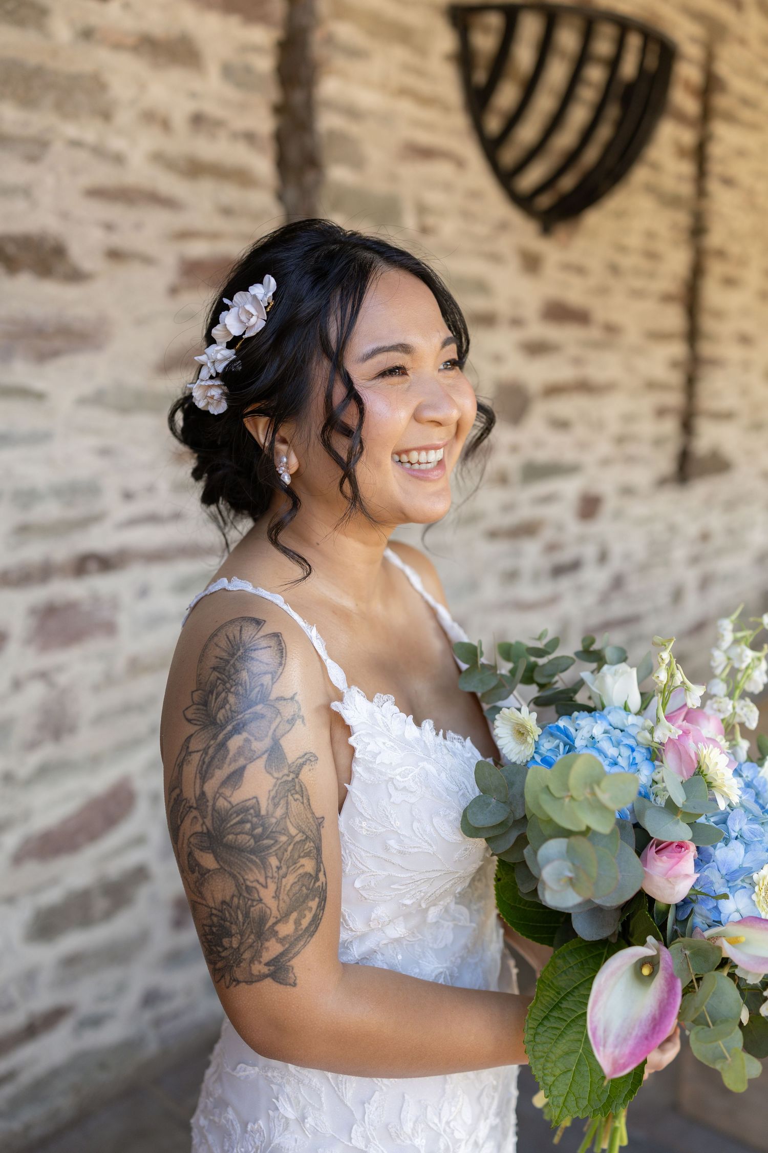 Smiling bride in white lace gown with floral hair accessories and arm tattoo holds blue and pink bouquet outdoors.