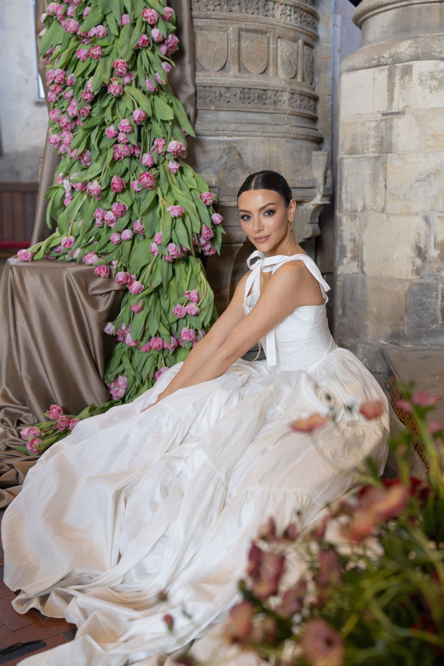 Elegant white wedding gown with flowing skirt photographed against stone columns and pink floral arrangements.