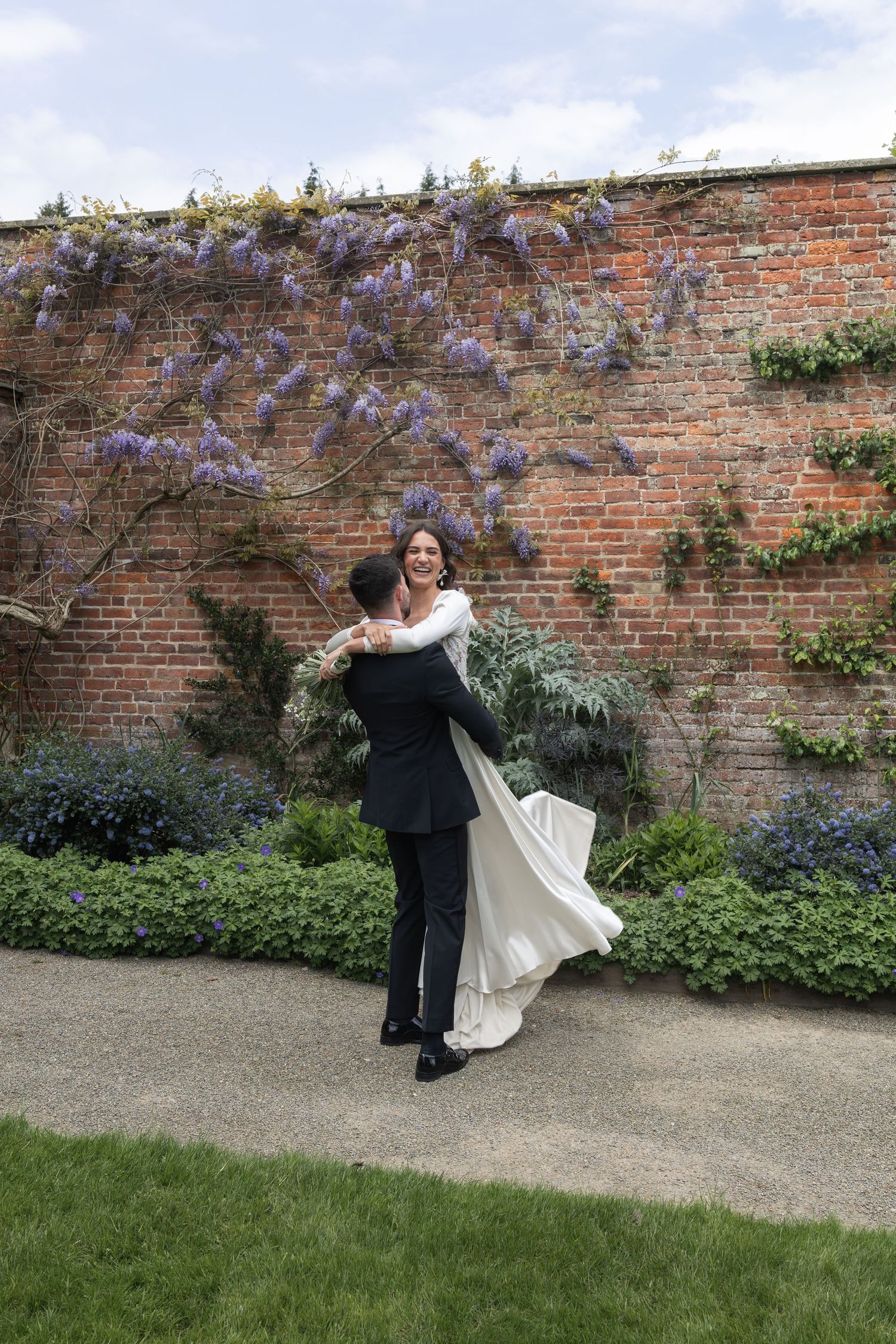 Bride and groom dancing in a walled garden with purple wisteria, groom lifting bride in white dress.