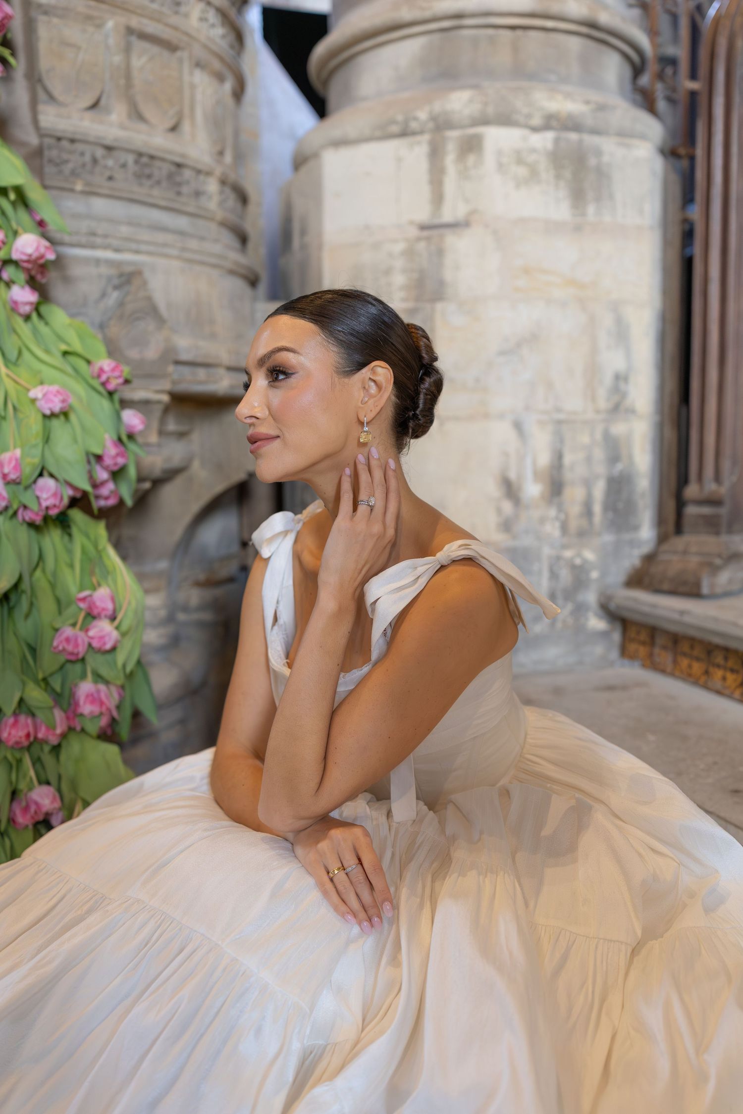 A bride in an elegant white ballgown poses gracefully against stone pillars with pink floral decorations.