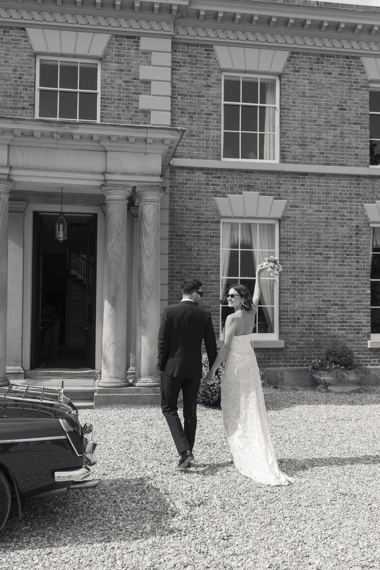 Black and white photo of bride and groom walking hand in hand toward a grand brick manor house with classic car nearby.